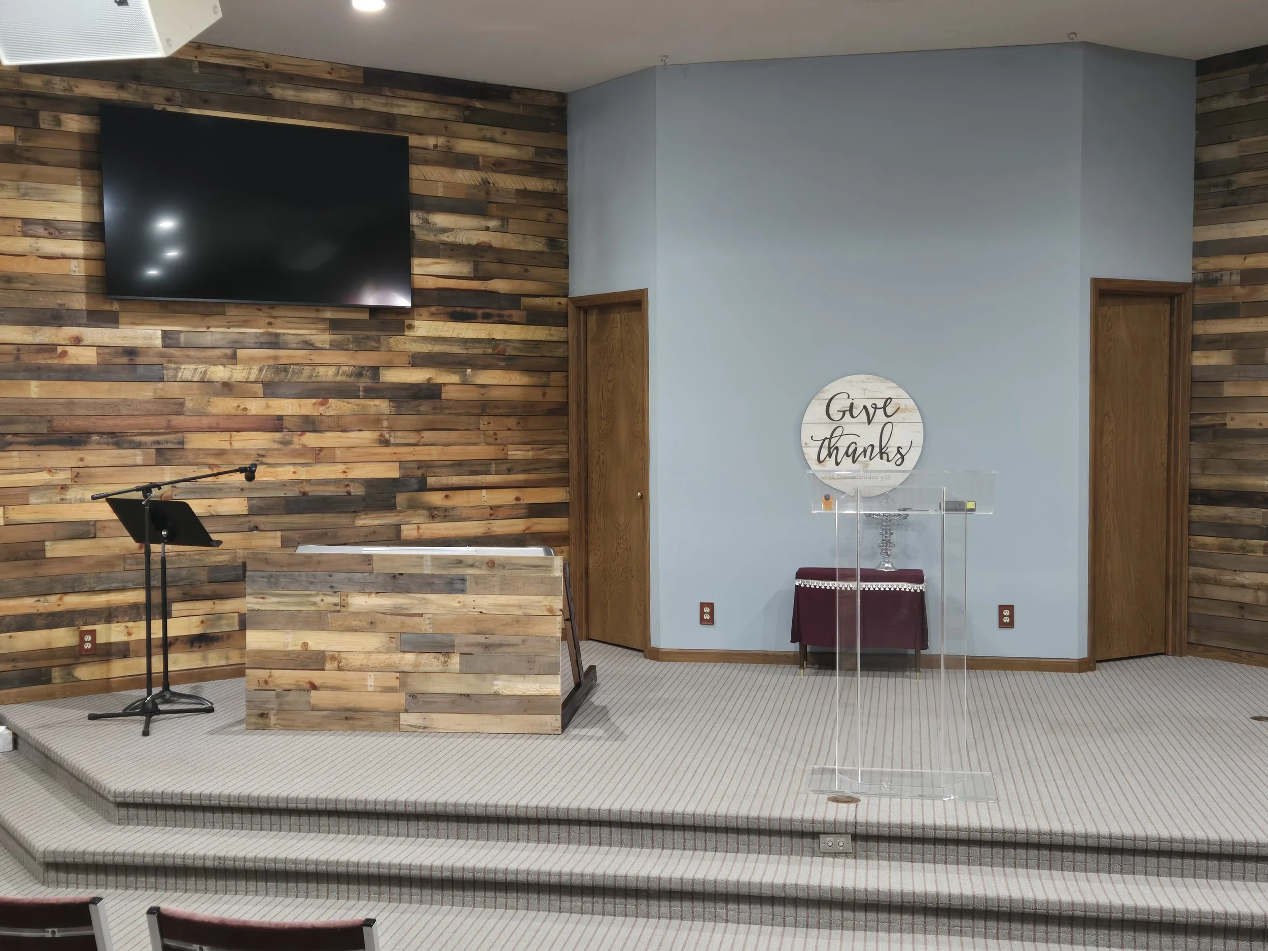 Church sanctuary stage with rustic wood-paneled walls, wall-mounted TV, lectern, microphone stand, and glass communion table beneath a “Give Thanks” sign.