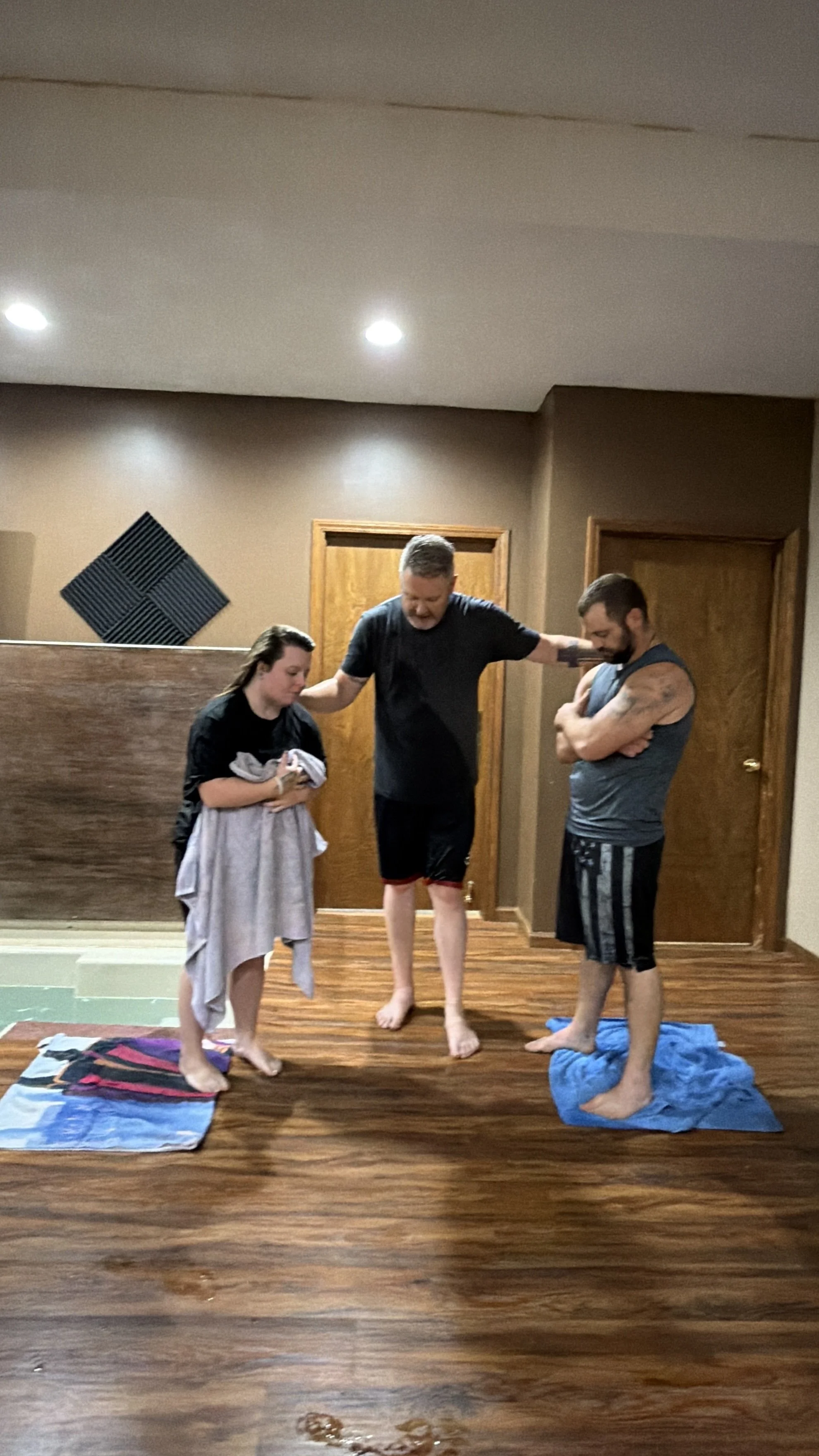 Three barefoot adults stand indoors on towels in a wood-paneled room, with one person holding a folded towel.