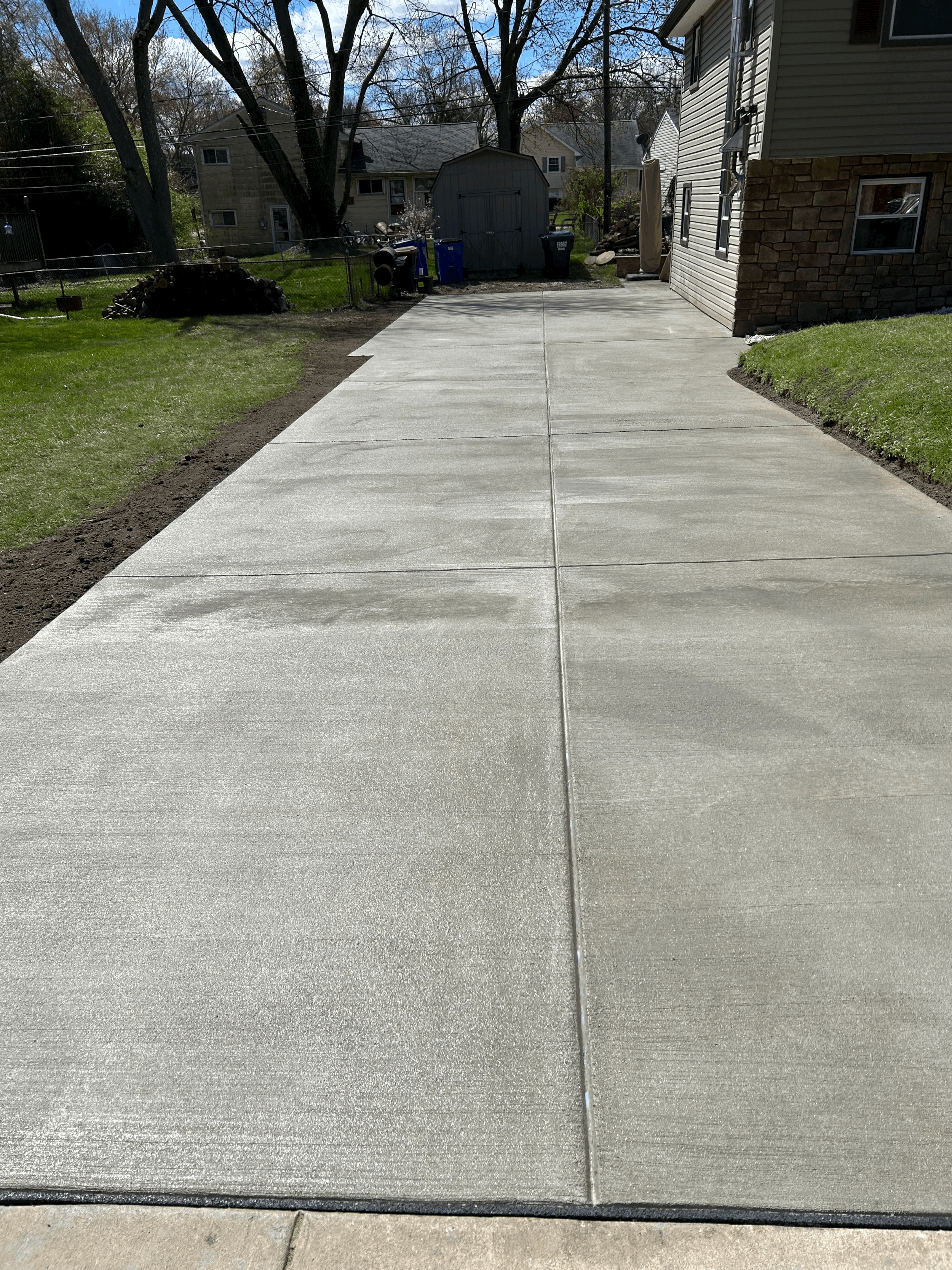 Newly poured concrete driveway in a residential backyard, bordered by grass and a house on the right, with trees and a shed in the background.
