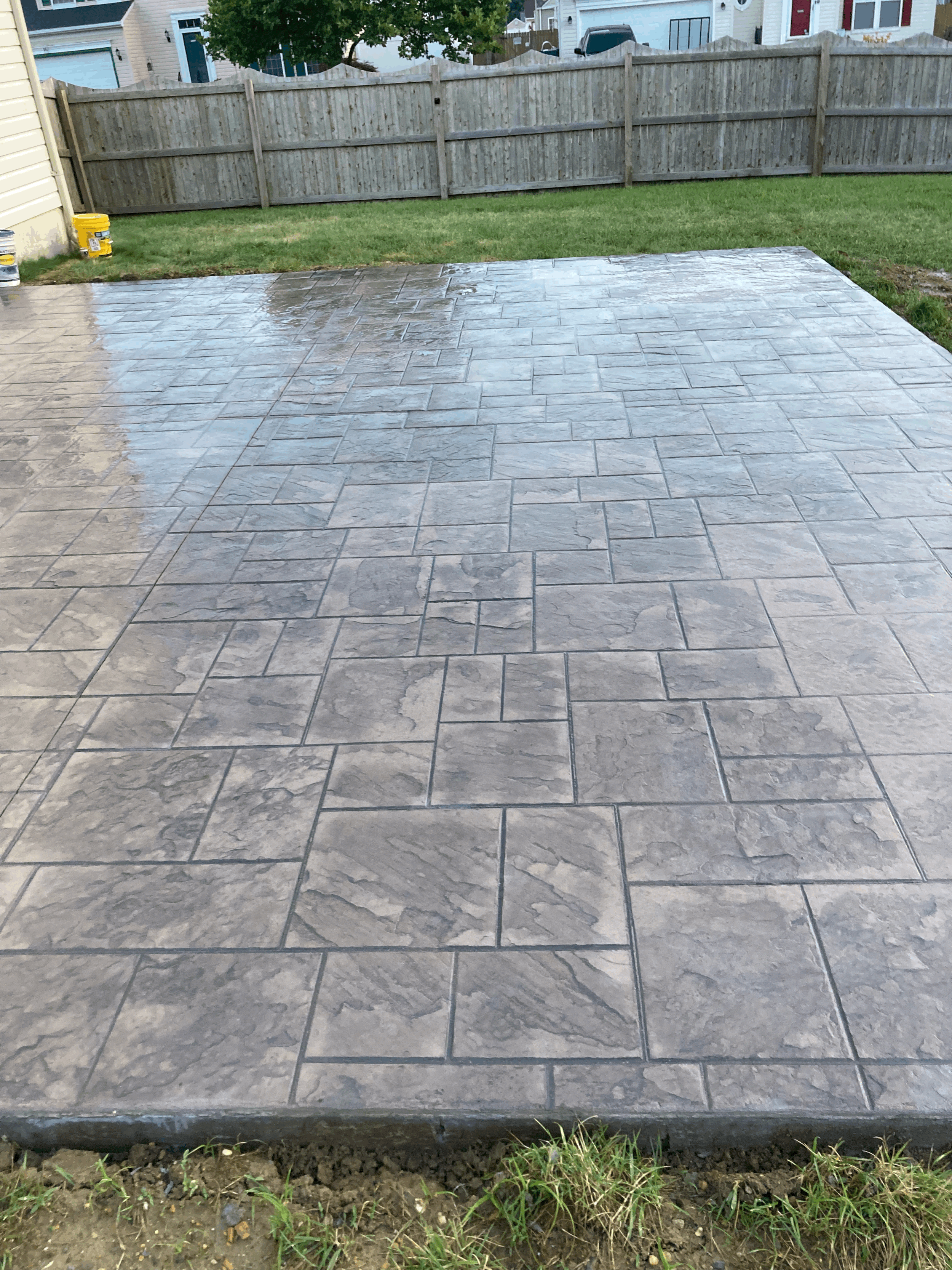 Freshly paved concrete patio with stamped stone pattern in a backyard, bordered by grass and a wooden fence.