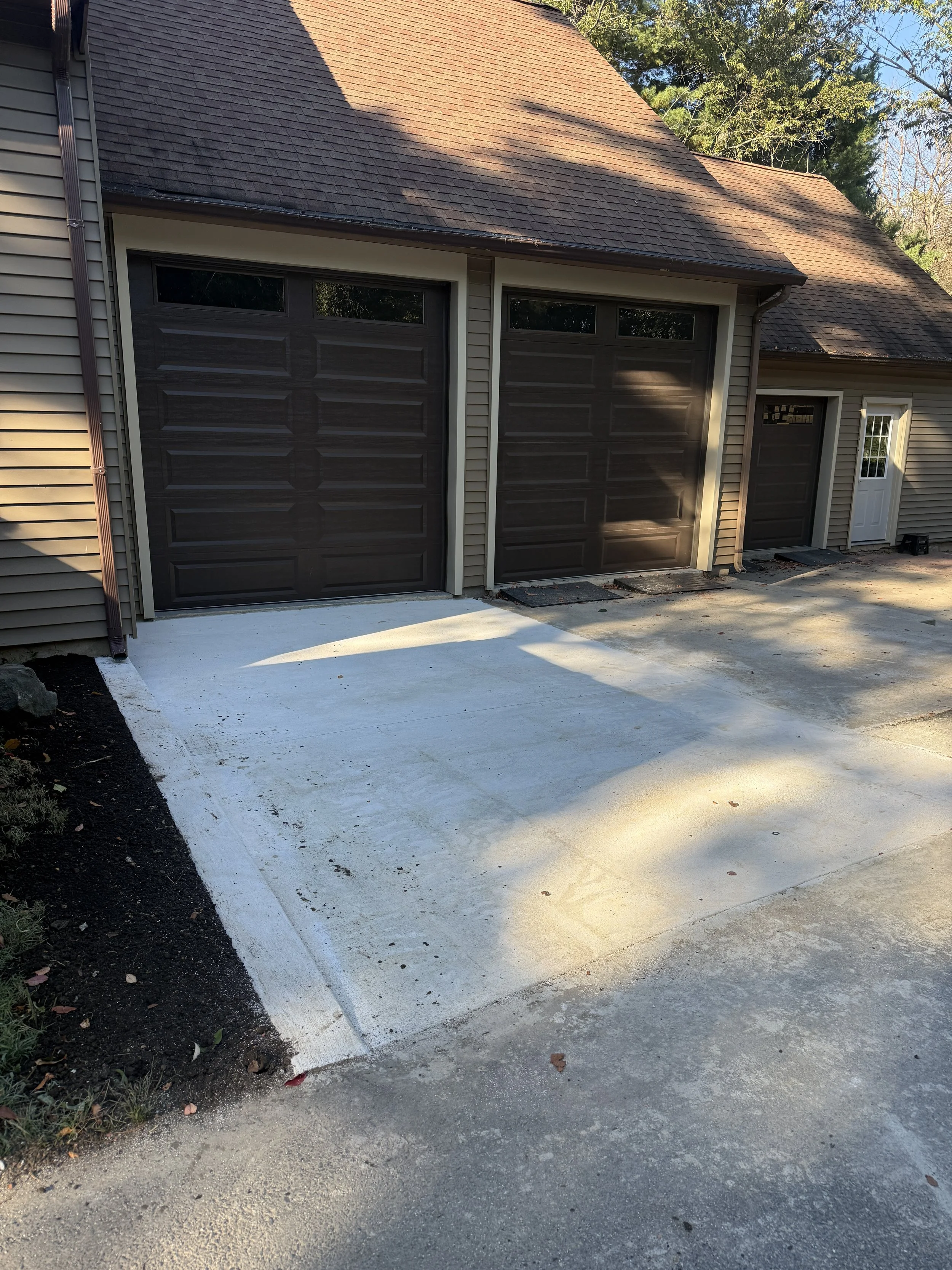 Newly poured concrete driveway in front of a house with two garage doors and a side door, surrounded by a garden bed on the left and a paved area on the right.