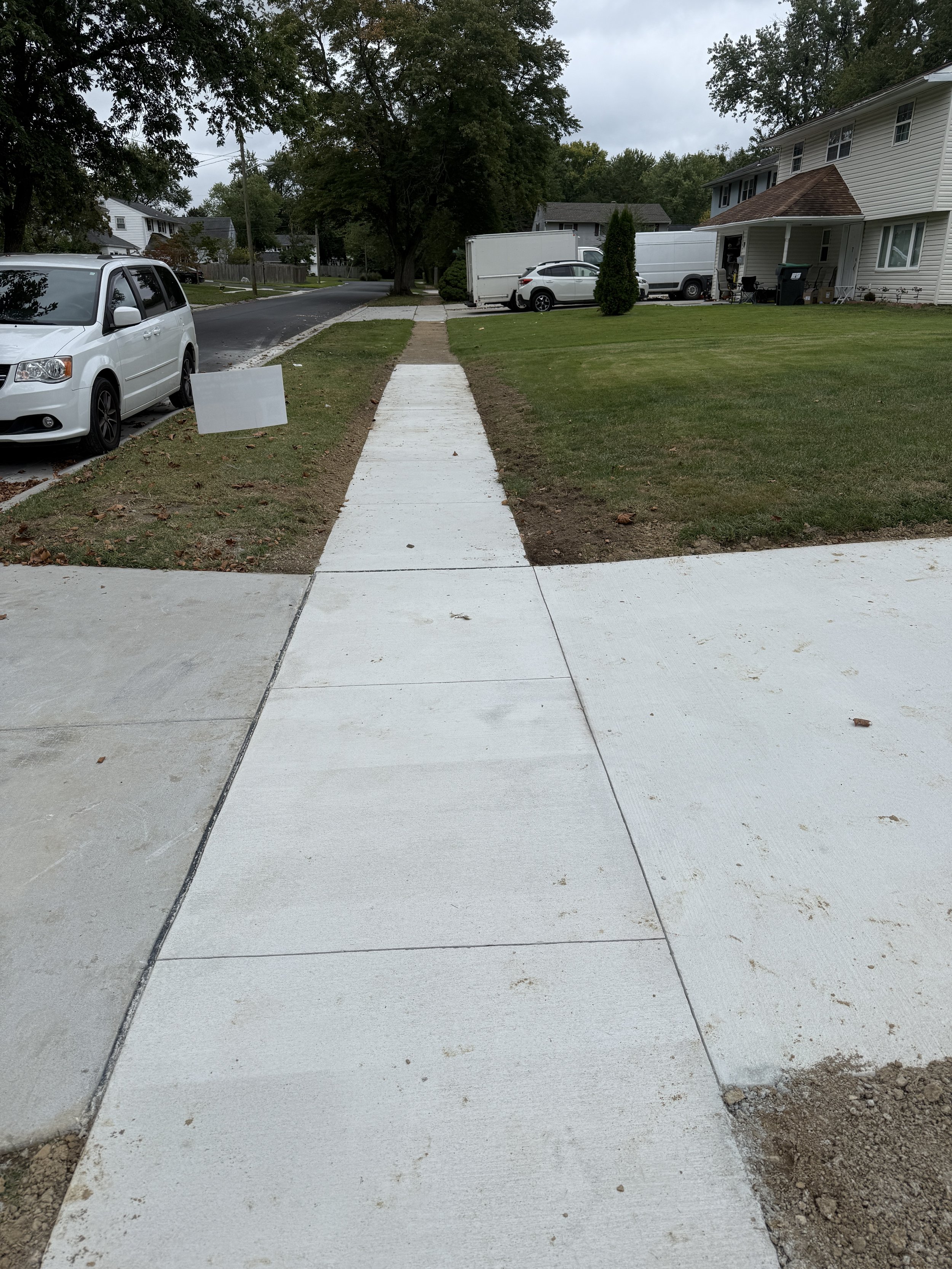 A freshly paved concrete sidewalk leading from the house to the street in a suburban neighborhood, with a grassy lawn on each side and parked cars nearby.
