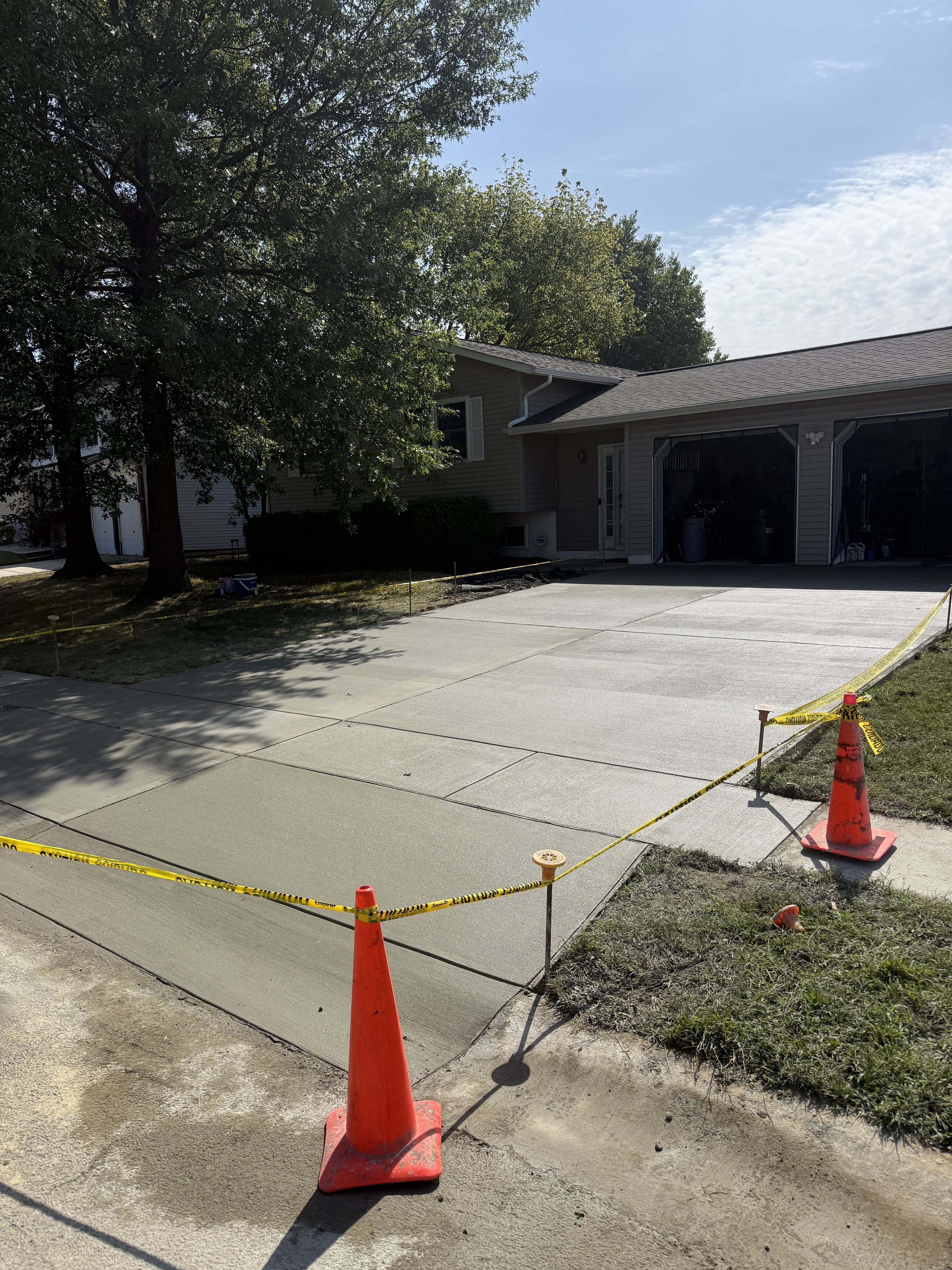 Newly poured concrete driveway section with orange cones and yellow caution tape, in front of a house with a two-car garage, trees, and partly cloudy sky.