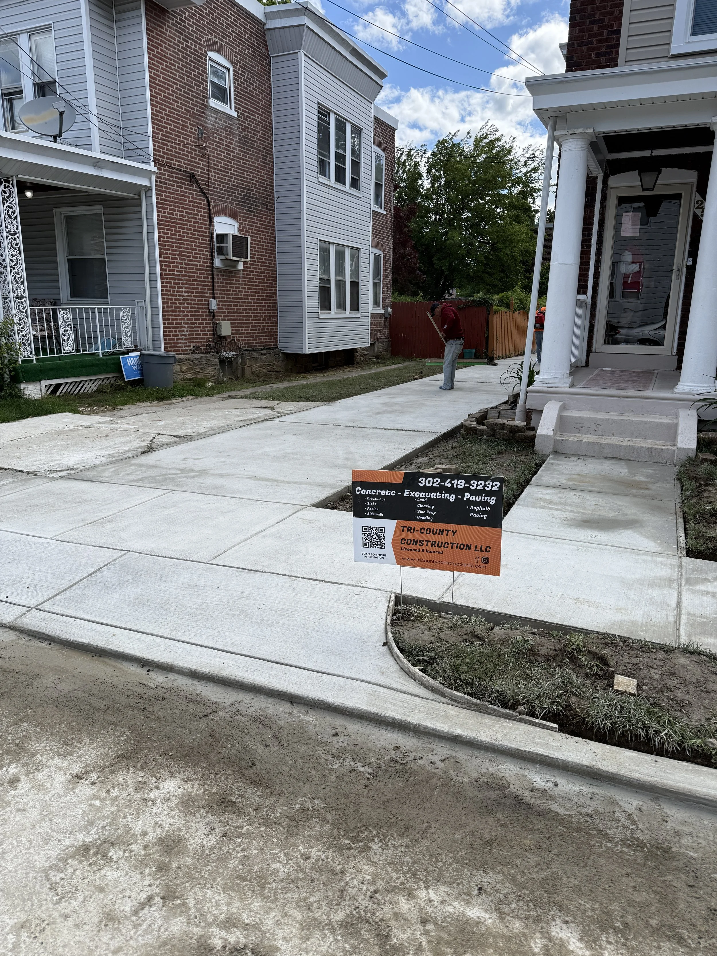 New concrete sidewalk in front of house with construction sign for Tri-County Construction LLC, showing concrete, excavating, and paving services. Worker is using a rake in the background.
