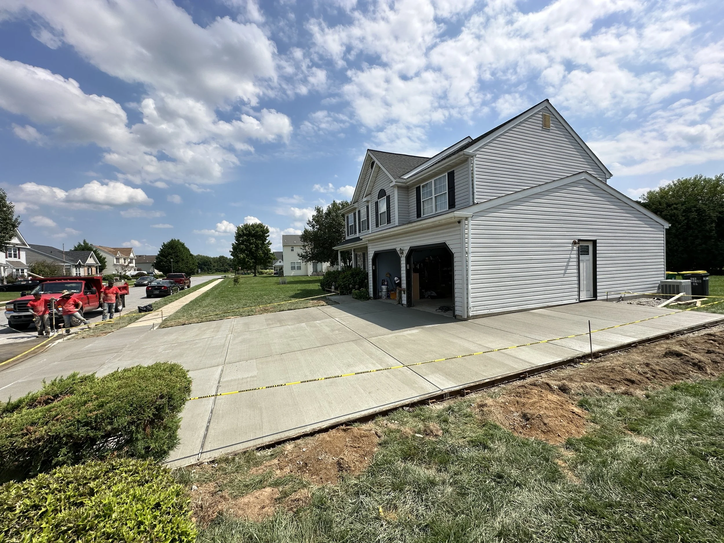 Newly poured concrete driveway in front of a two-story house with a gray exterior, garage door open, and construction crew working on the side. Residential neighborhood with other houses, cars, trees, and a clear blue sky.