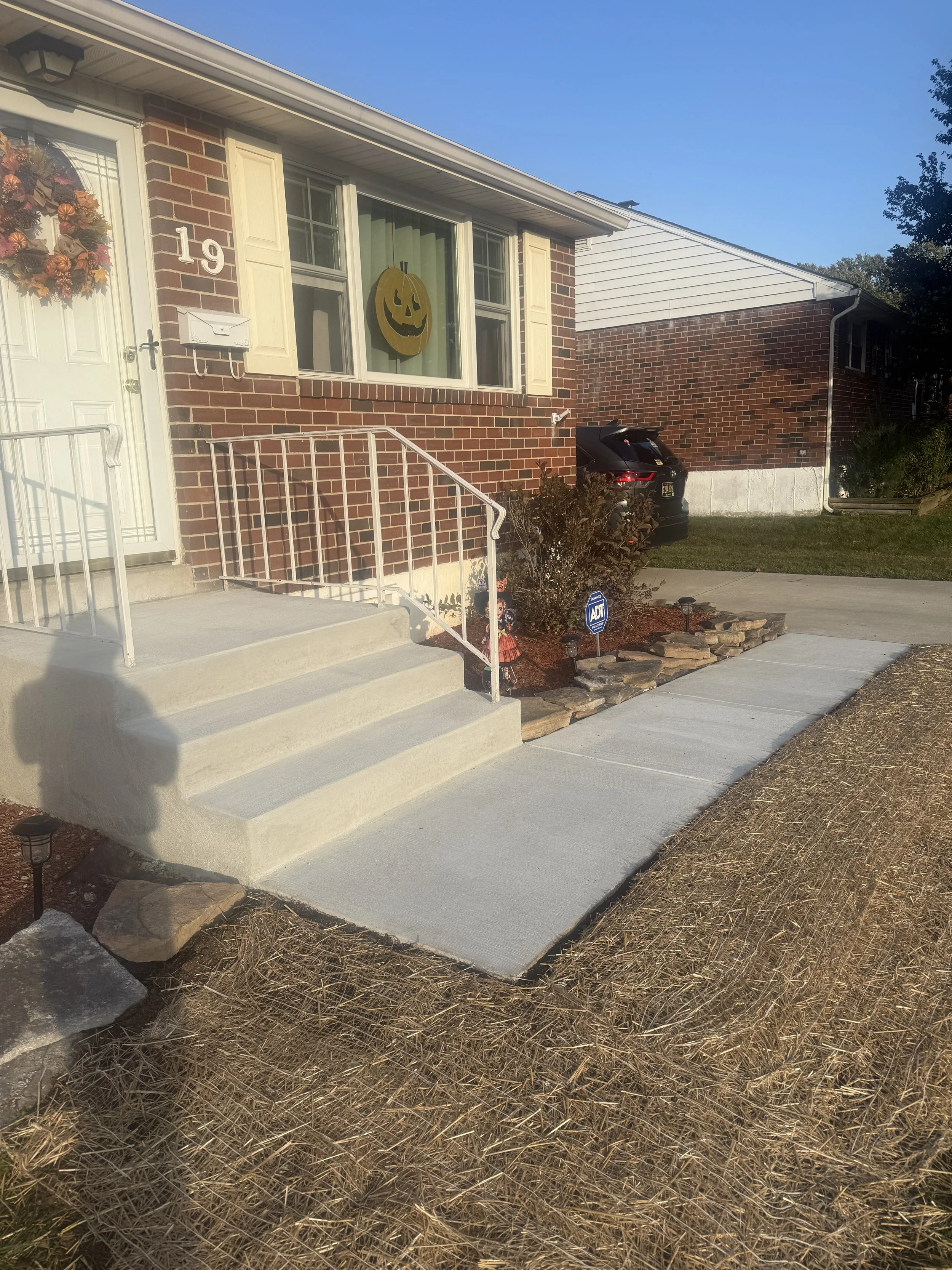 Front porch of a brick house with a concrete walkway and steps, Halloween decorations including a pumpkin sign in the window, a wreath on the door, and an ADT security sign in the garden.