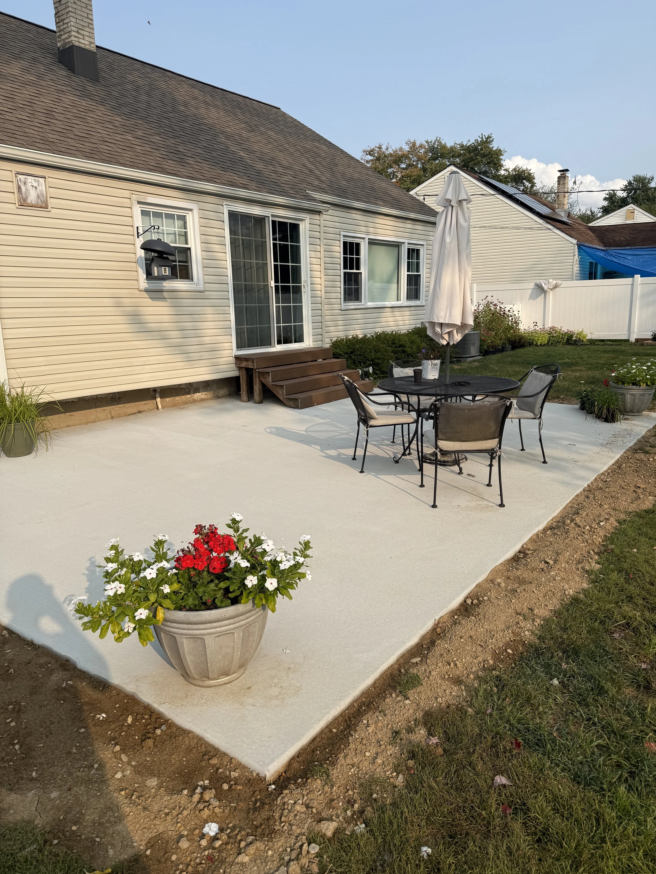 A backyard patio with a round black table, six chairs, a white patio umbrella, potted flowers, and a sliding glass door leading to a house with beige siding.