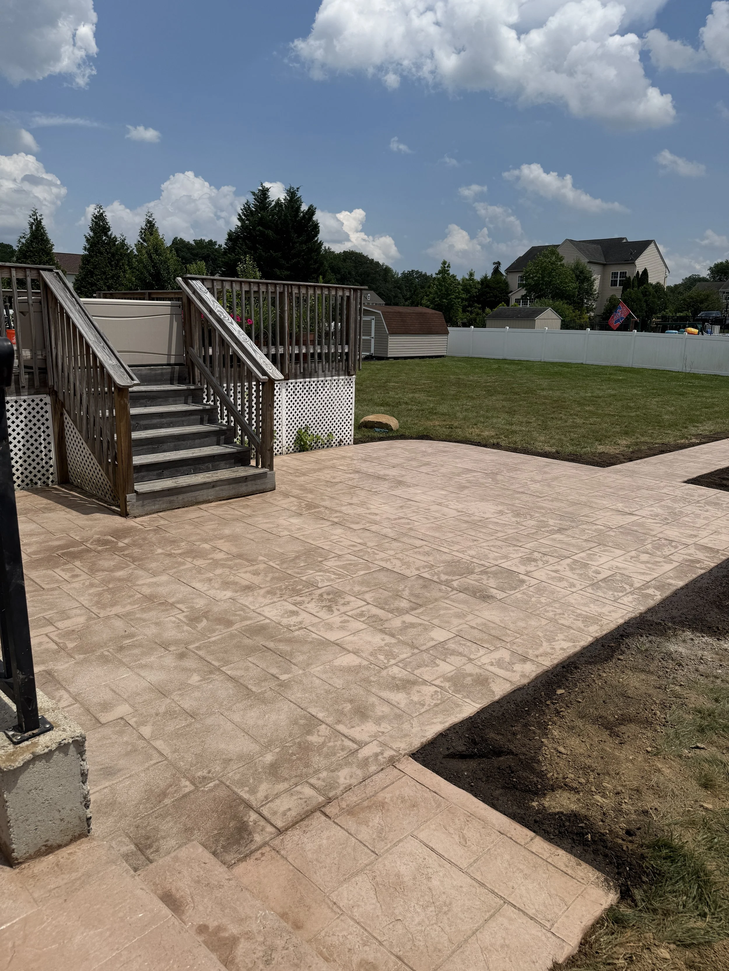 A backyard with a newly installed patio, wooden stairs leading to a deck, a grassy lawn, and houses in the background under a partly cloudy sky.