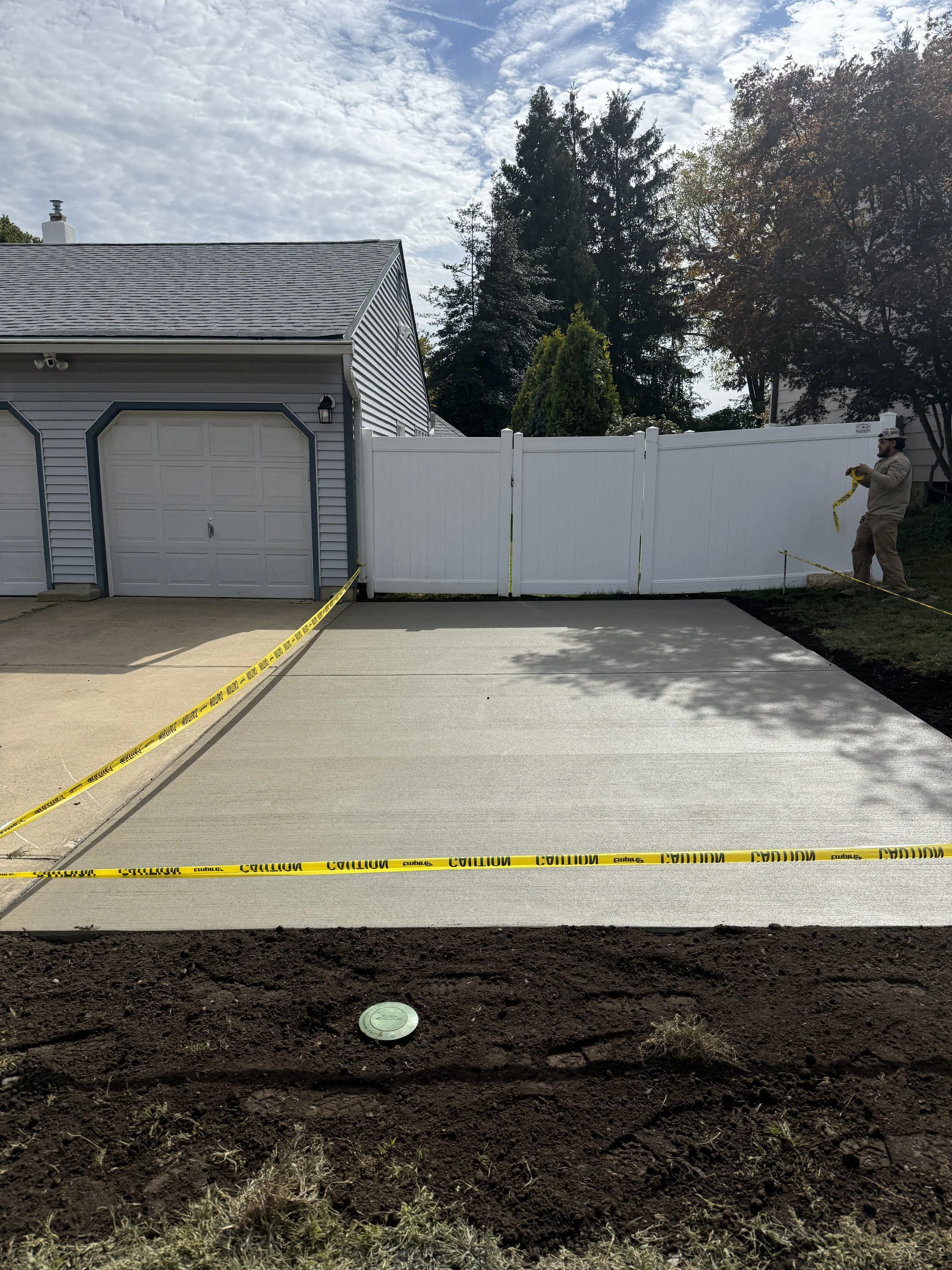 Newly poured concrete driveway section fenced off with yellow caution tape, adjacent to a white vinyl fence, with a man measuring on the right side, in front of a blue garage and house, under a partly cloudy sky.