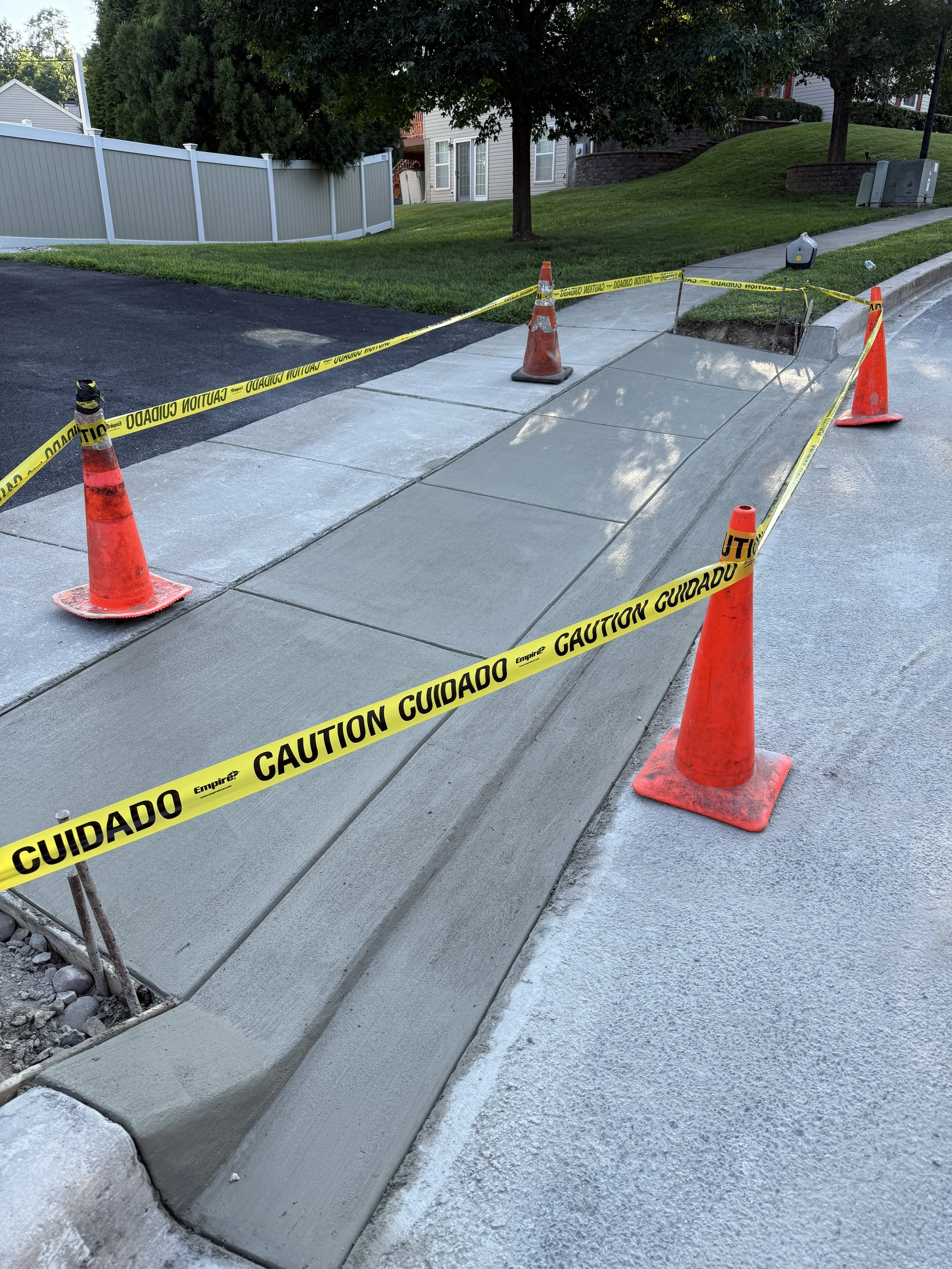 Newly poured concrete sidewalk section with caution tape and orange traffic cones around it, indicating ongoing construction or curing process.