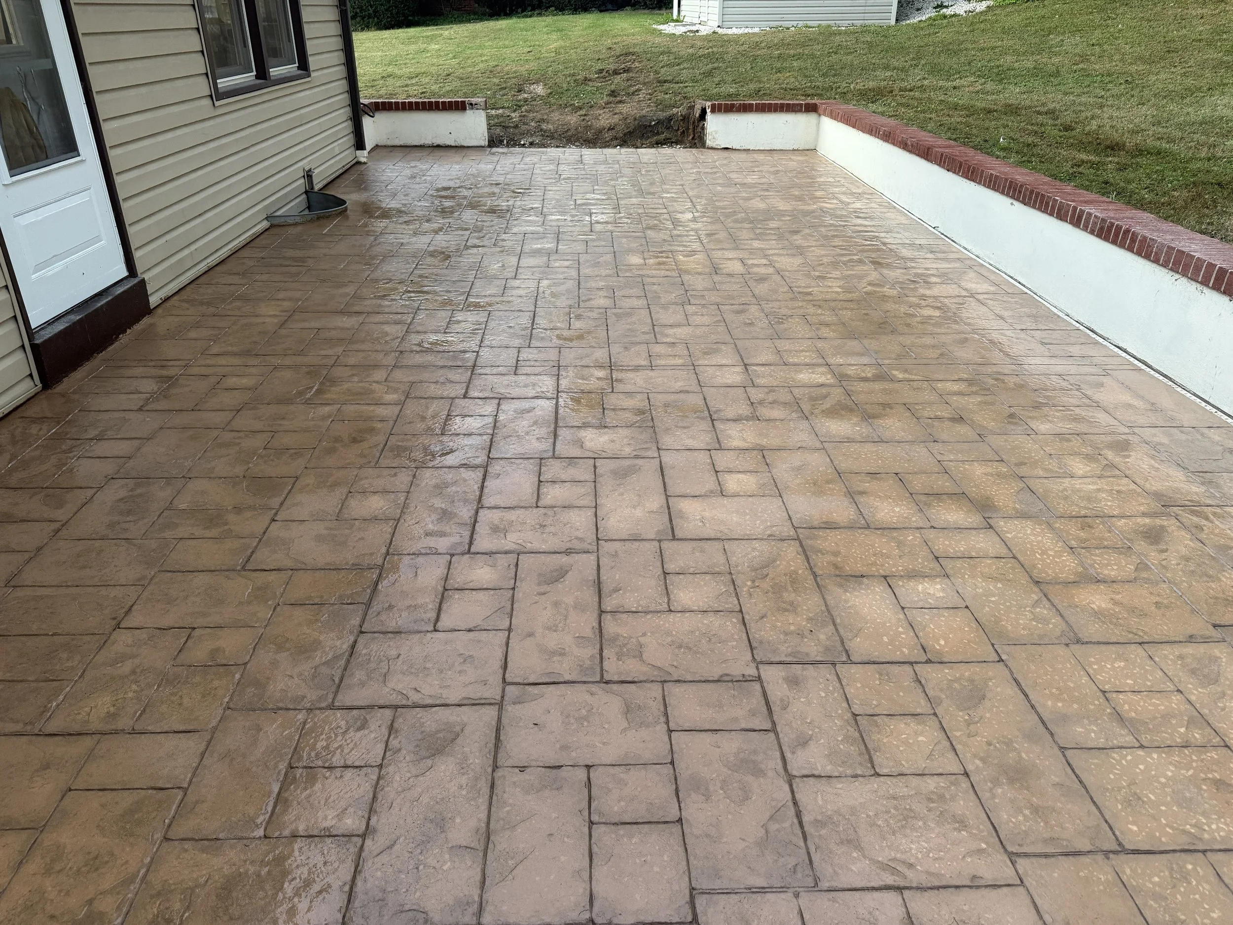 Wet stamped concrete patio area outside a house, with a brick and white wall border, and a grassy lawn beyond.