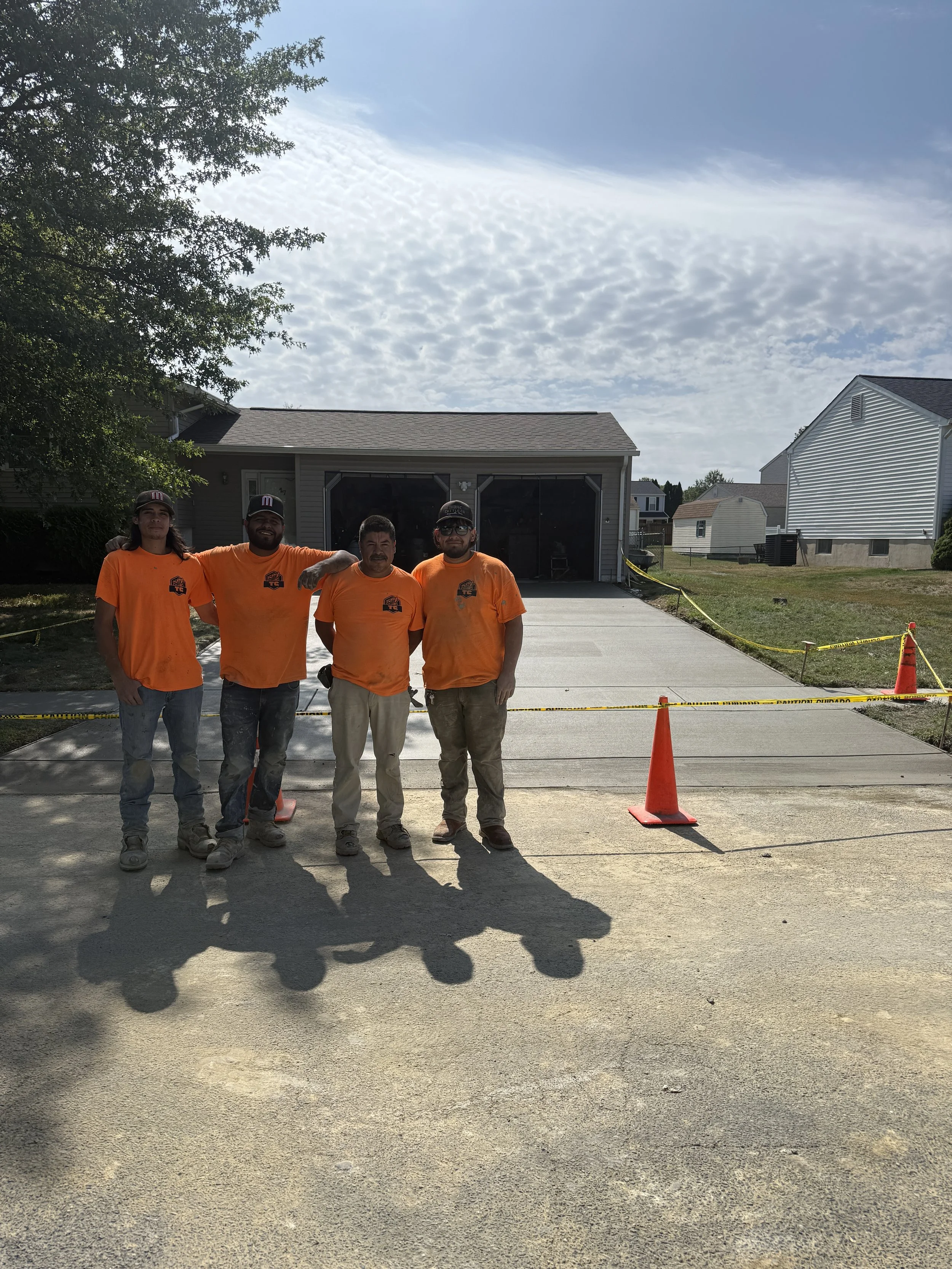 Four construction workers in orange shirts standing in front of a newly poured concrete driveway, with a house and neighboring homes in the background, yellow caution tape and orange cones surrounding the driveway.