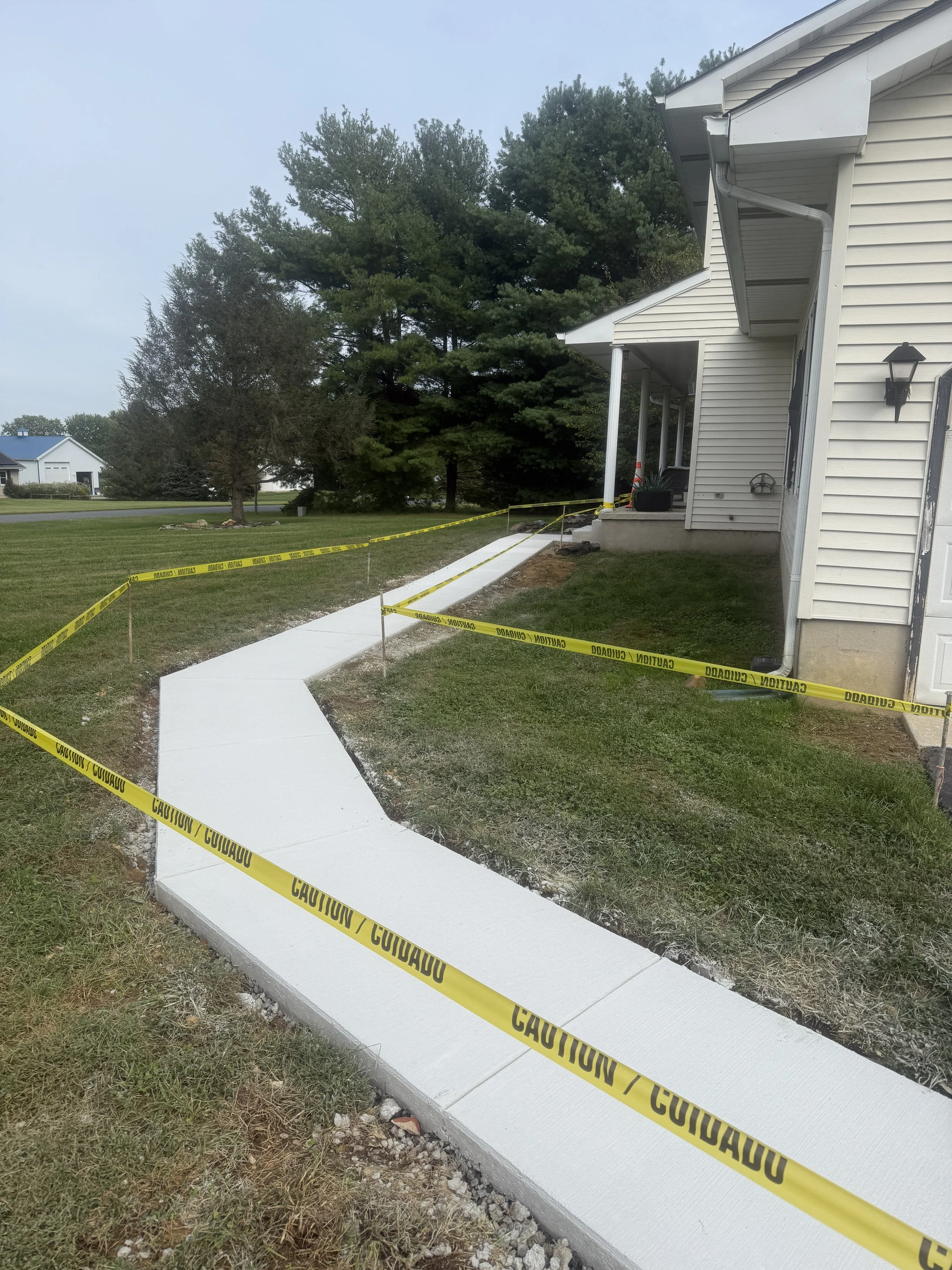 Newly paved sidewalk with caution tape around it in front of a house with white siding, porch, and outdoor wall-mounted lantern, surrounded by grass and trees.