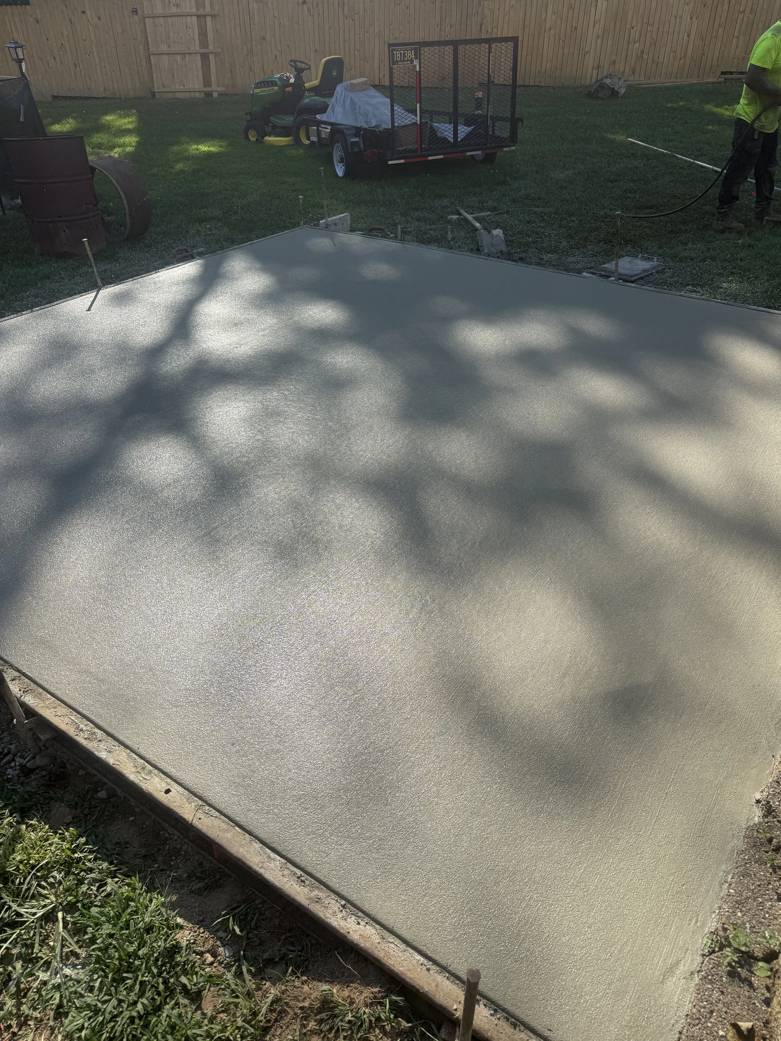 Freshly poured concrete slab in a backyard, with construction stakes and tools around it, and a worker spraying water on the surface.