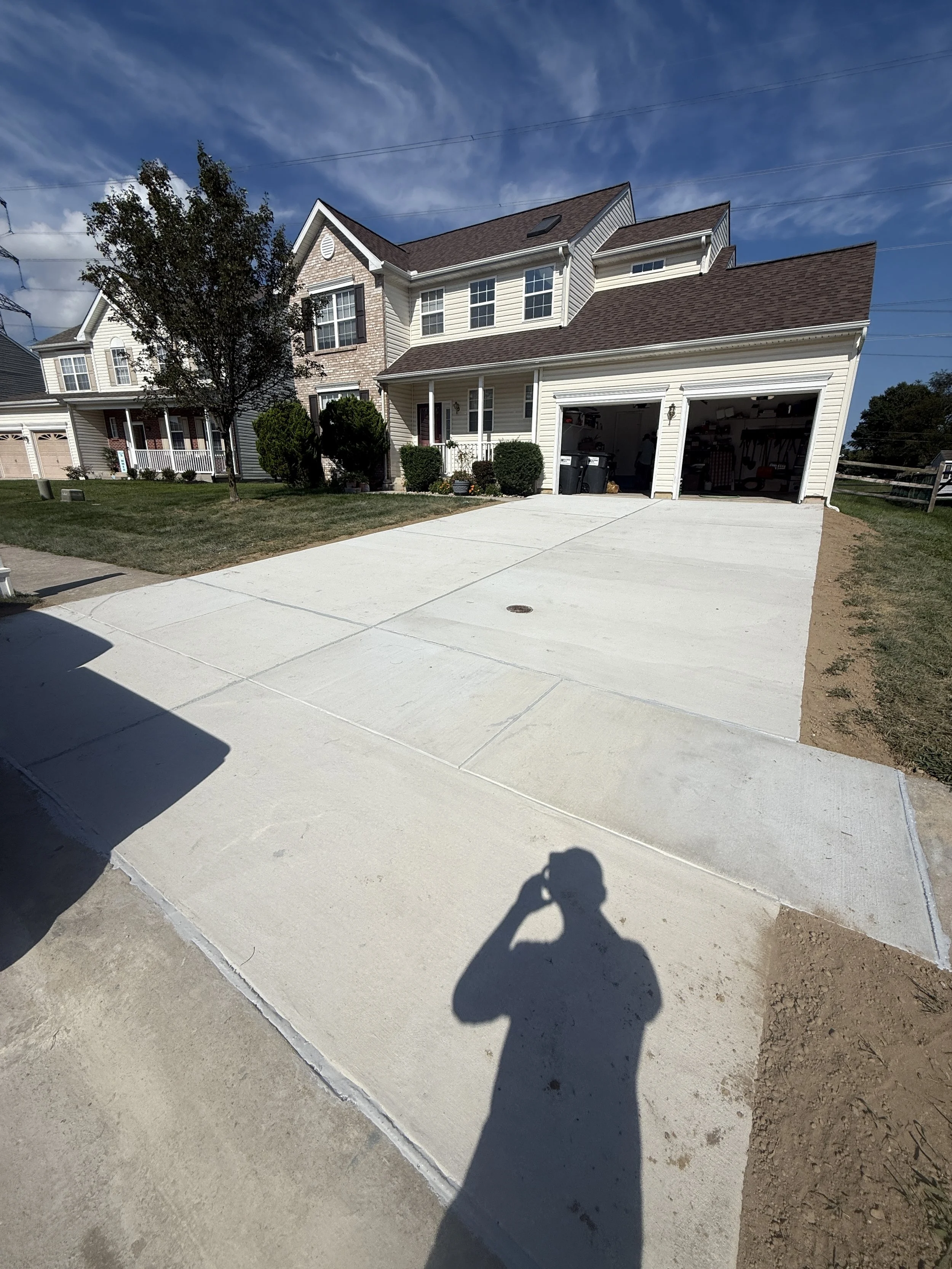 Newly poured concrete driveway in front of a two-story house with a garage, surrounded by a lawn and some bushes, with a person’s shadow taking the photo.