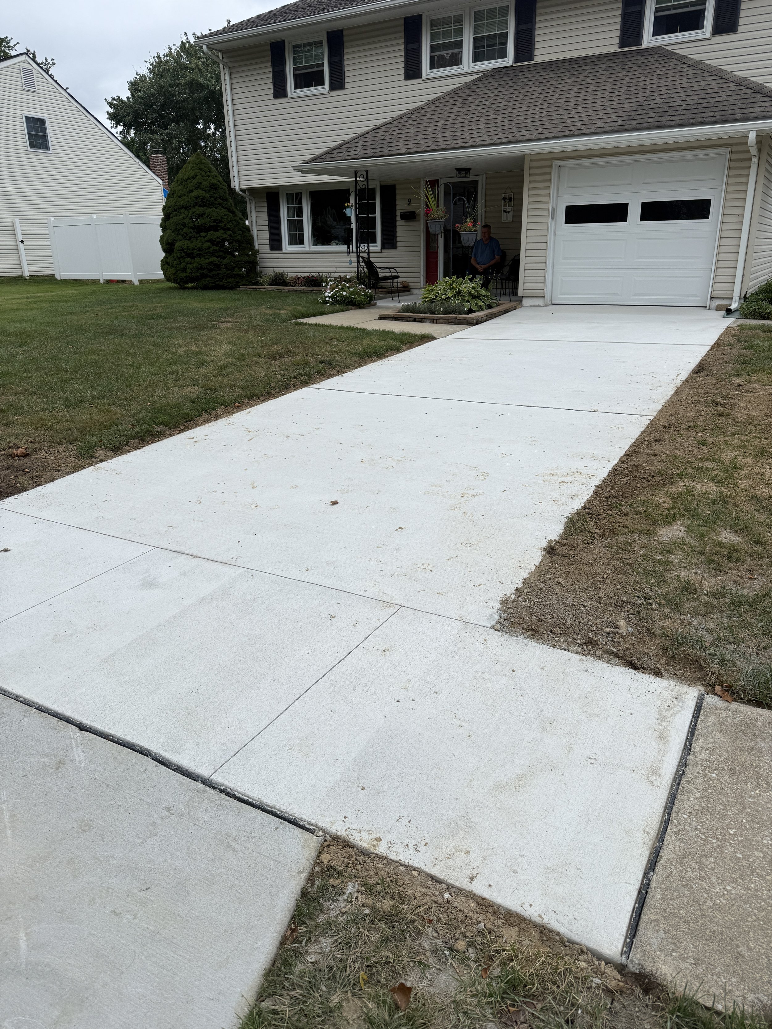 Newly paved concrete driveway leading to a house with a garage, front porch, and a man sitting outside on a bench. The lawn surrounding the driveway is partially patchy, with some grass and dirt.