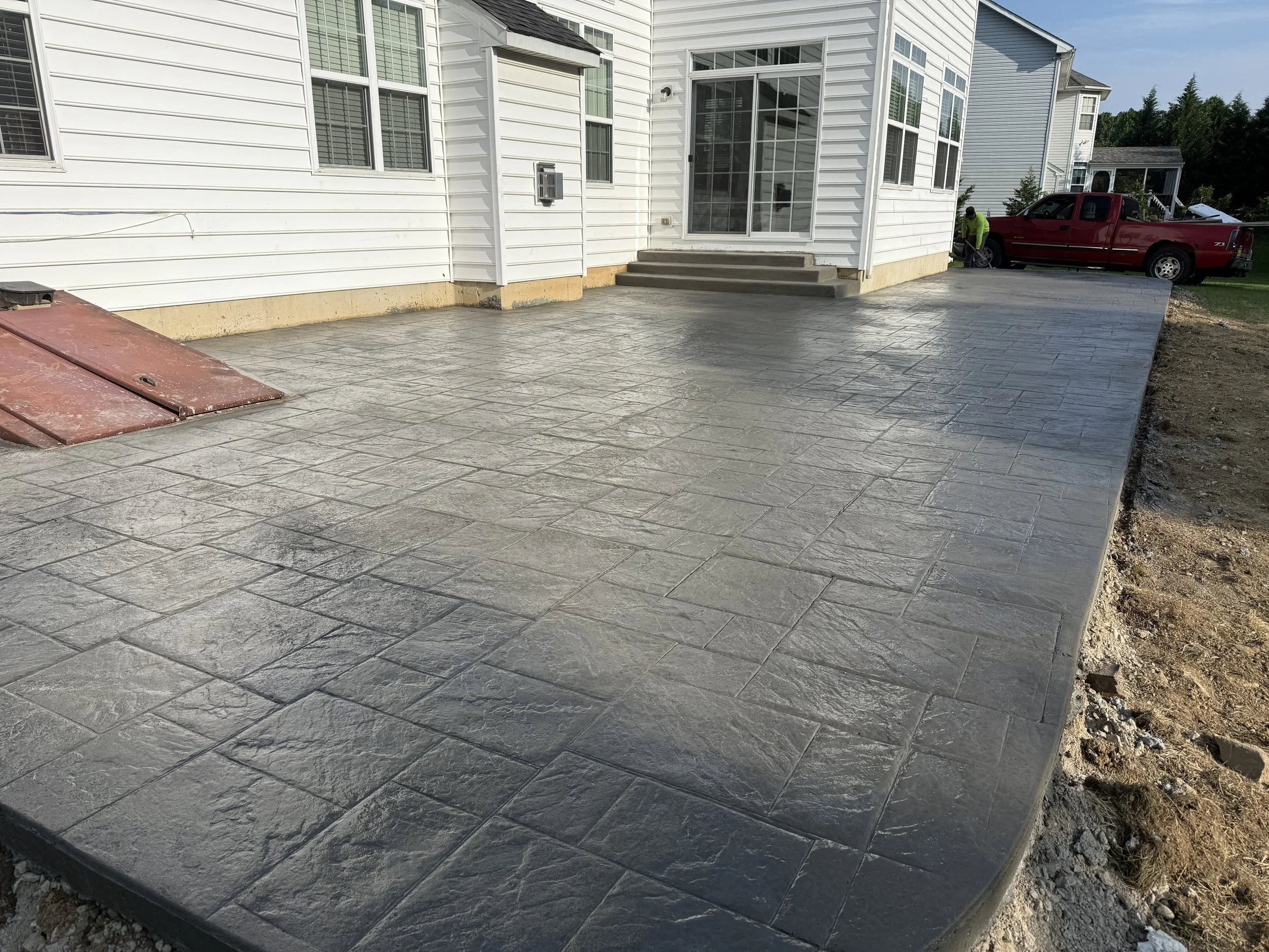 Freshly poured concrete patio with stamped pattern in front of a white house, with construction workers and a red truck in the background.