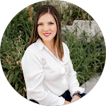 A young woman with long brown hair and a white blouse sitting outdoors among green plants and foliage, smiling at the camera.