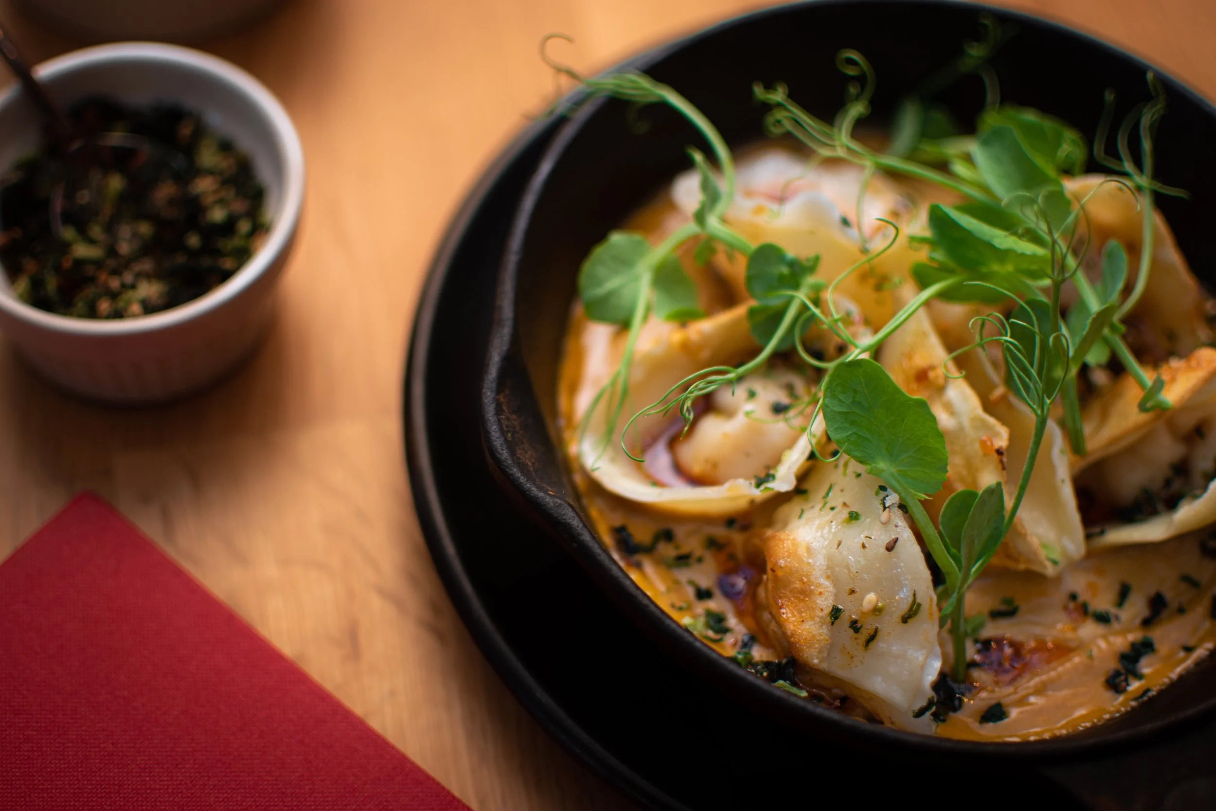 Gourmet dumplings in a black bowl garnished with fresh greens and herbs on a wooden table with a small cup of seasoning.
