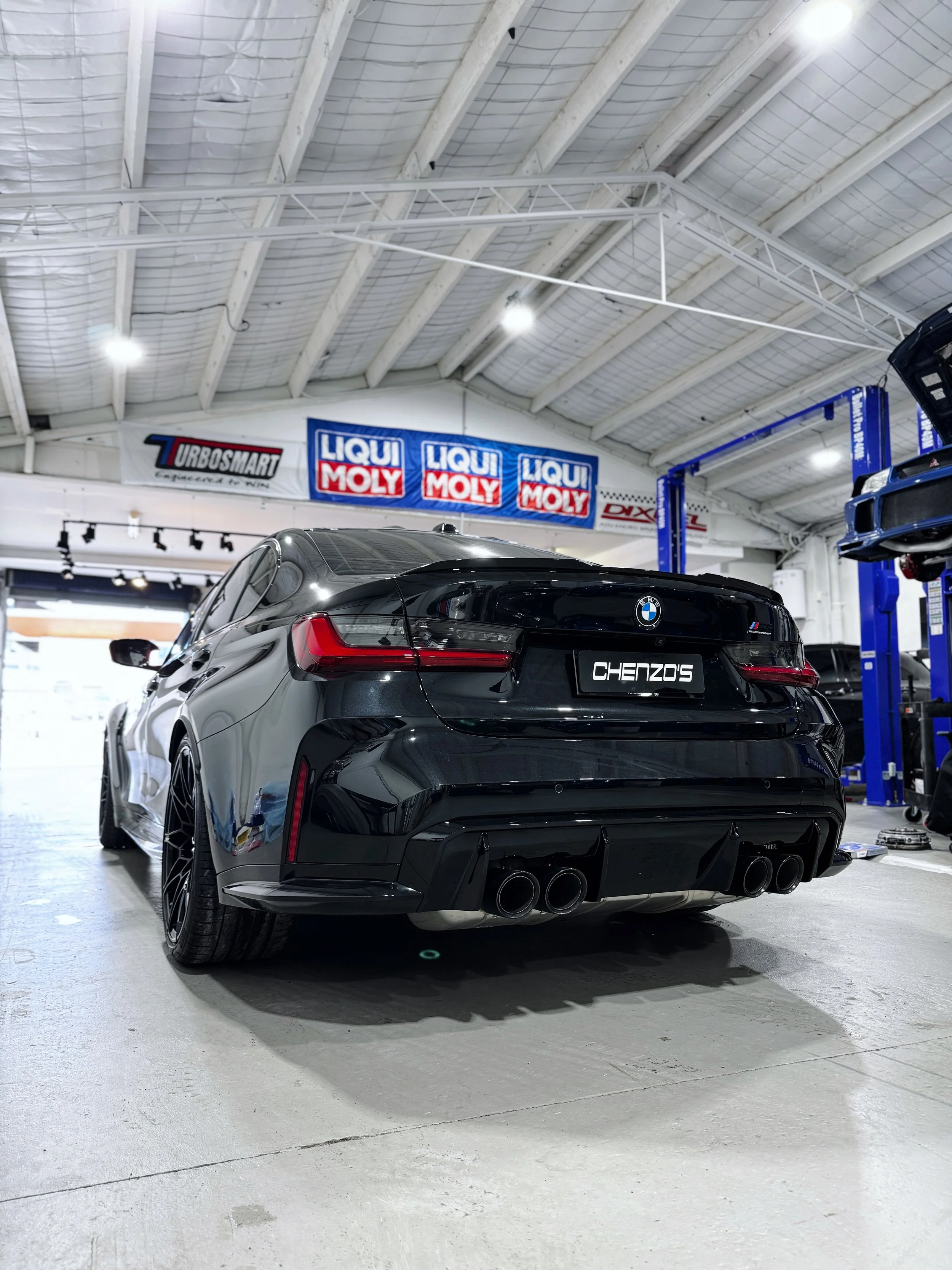 Black BMW M3 parked inside a garage or workshop with racing tires and tools, with banners for Liqui Moly and Urbosmart on the wall.