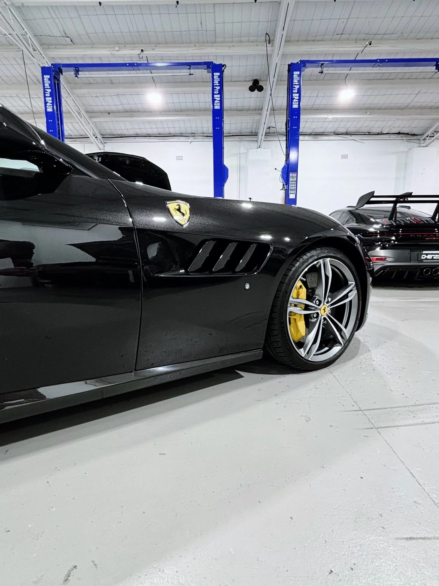 Close-up of a black Ferrari sports car with yellow brake calipers in a garage, showing the front side and wheel, with a Porsche in the background.