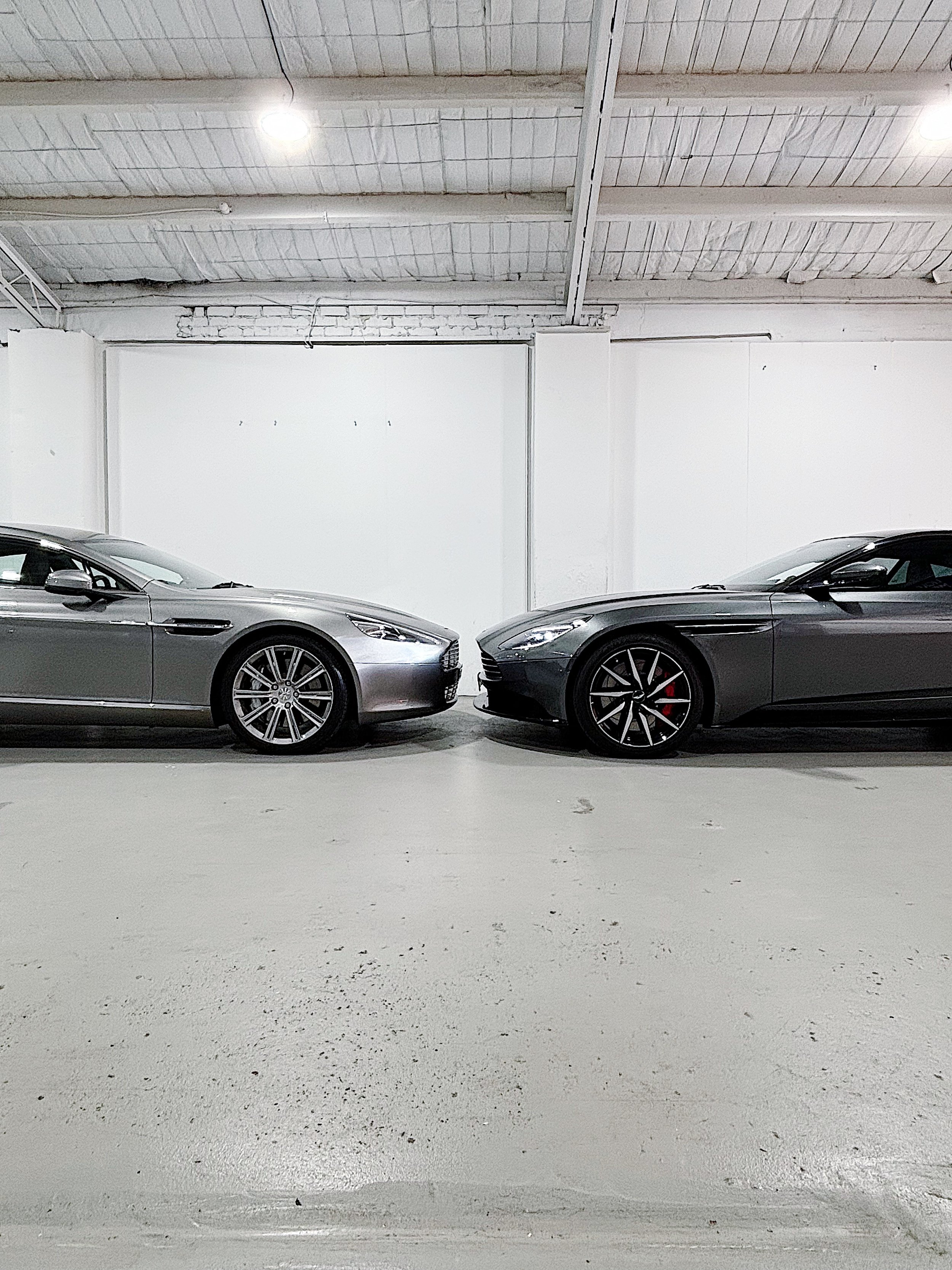 Two gray sports cars parked facing each other inside a concrete garage with white walls and industrial ceiling lighting.