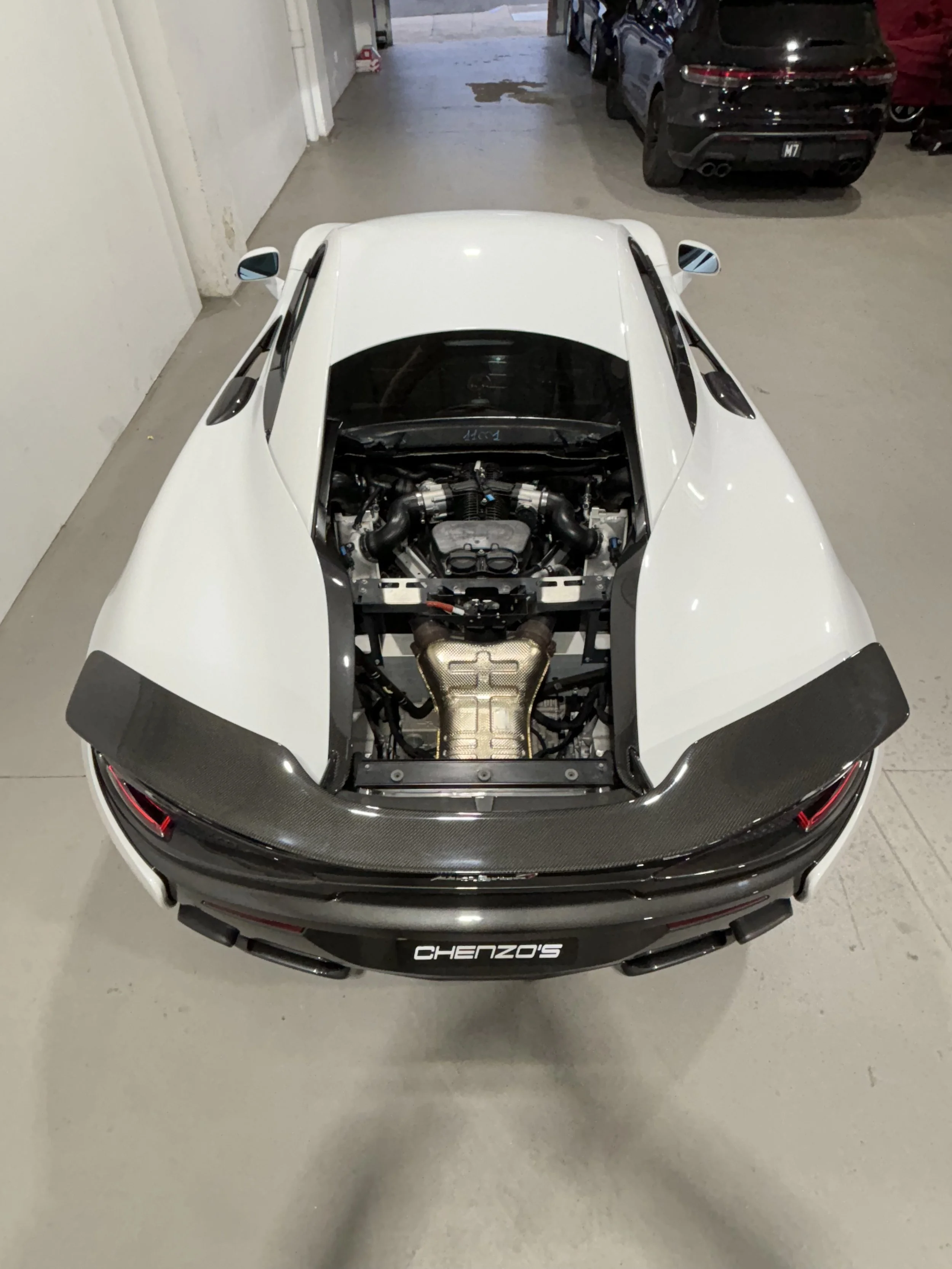 Rear view of a white sports car in a garage, showing the exposed engine and carbon fiber spoiler with a plaque that reads 'CHENZO'S'.