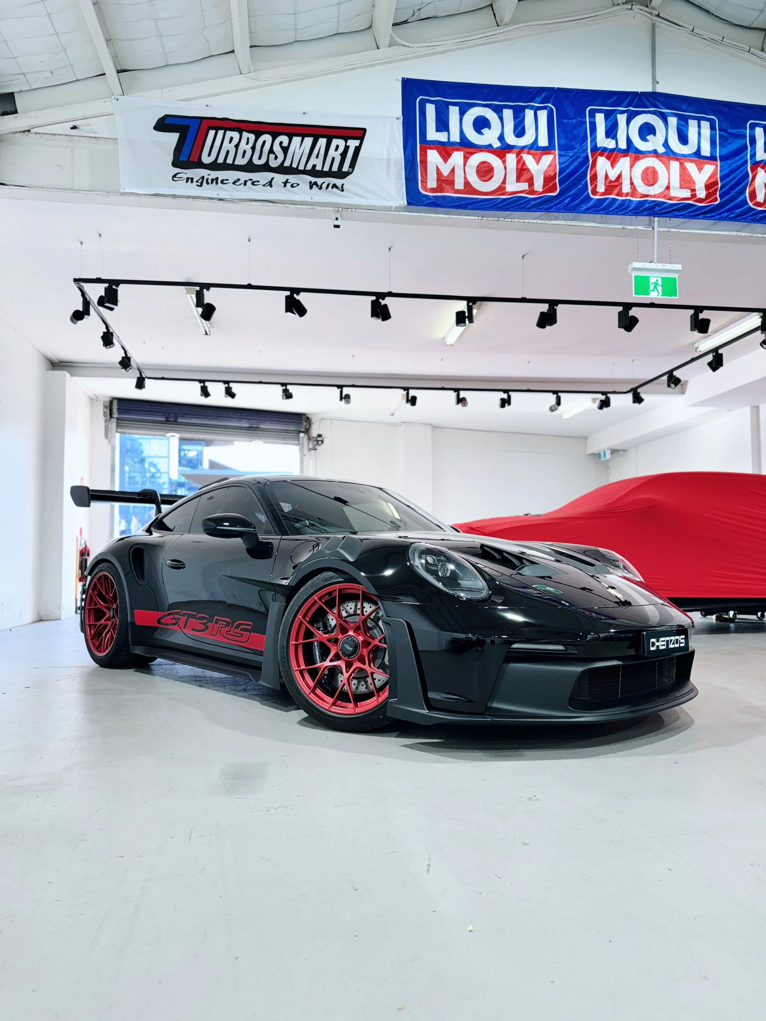 Black Toyota Supra GT 3 RS sports car with red wheels, parked indoors in a showroom with banners for TurboSmart and Liqui Moly on the ceiling.