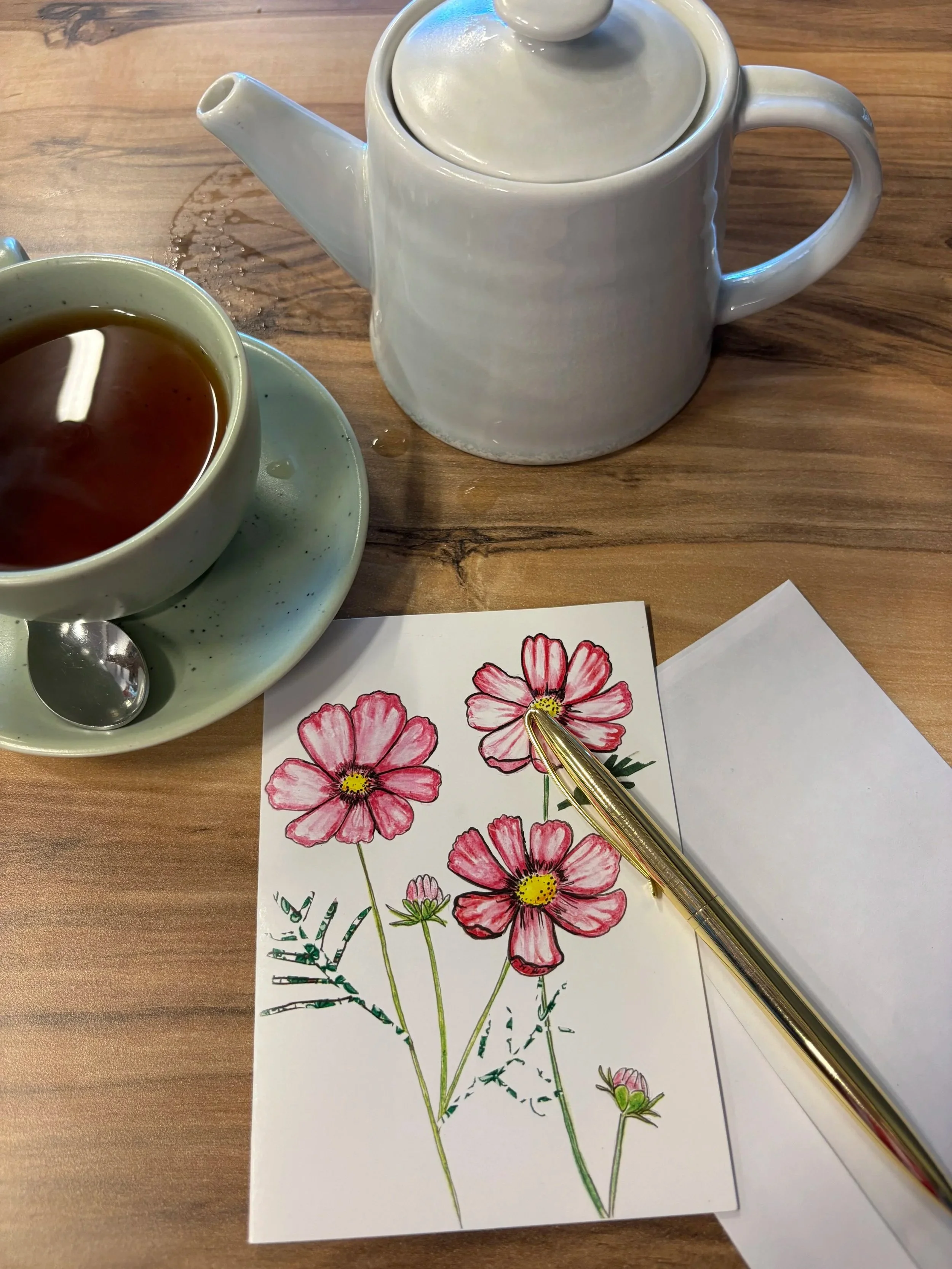 A teapot, a cup of tea on a saucer with a spoon, and a drawing of pink flowers with green stems and leaves on a wooden table.