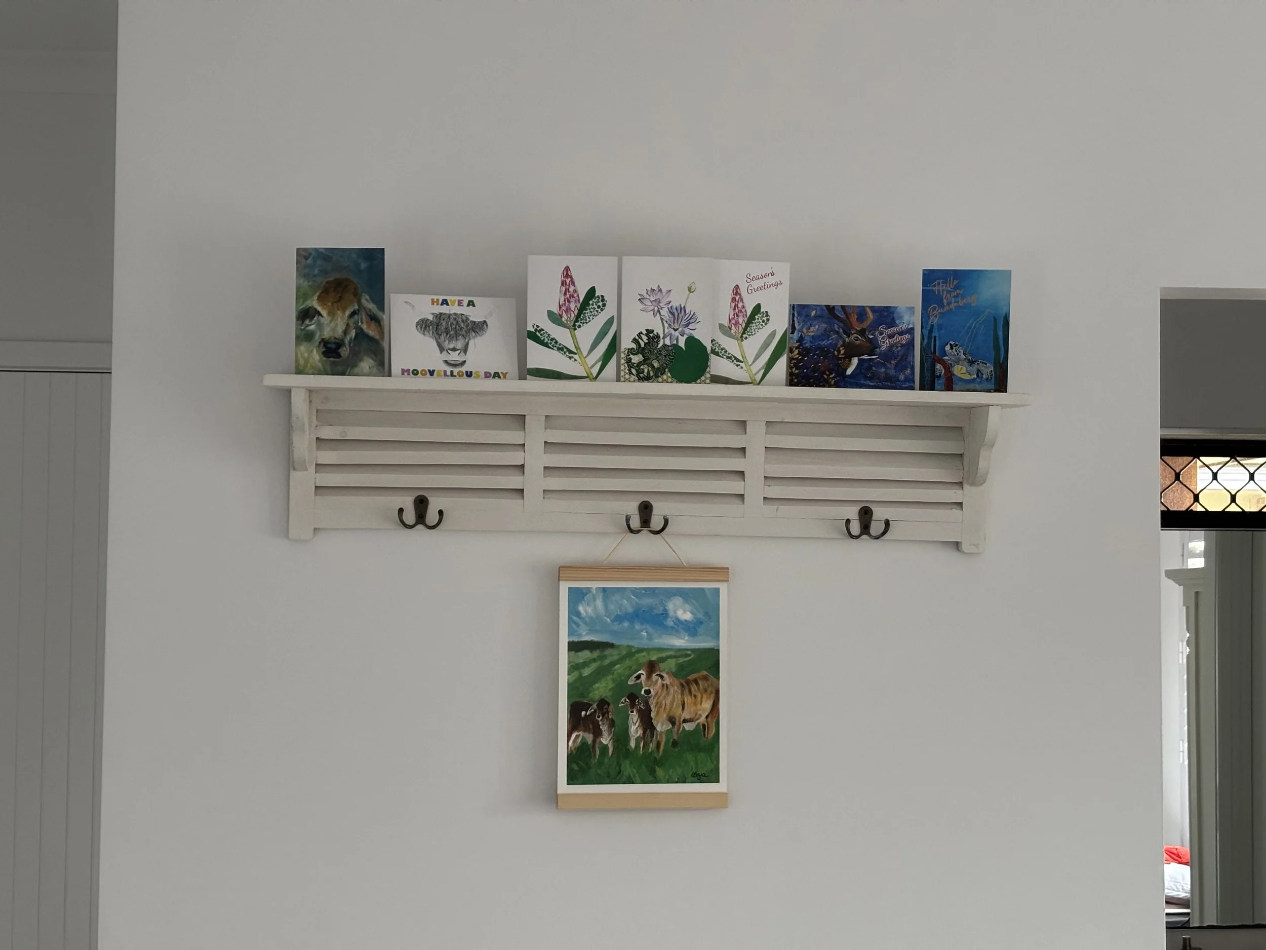 A white wall with a rustic wall shelf holding seven greeting cards, and a framed painting of cows hanging below the shelf.