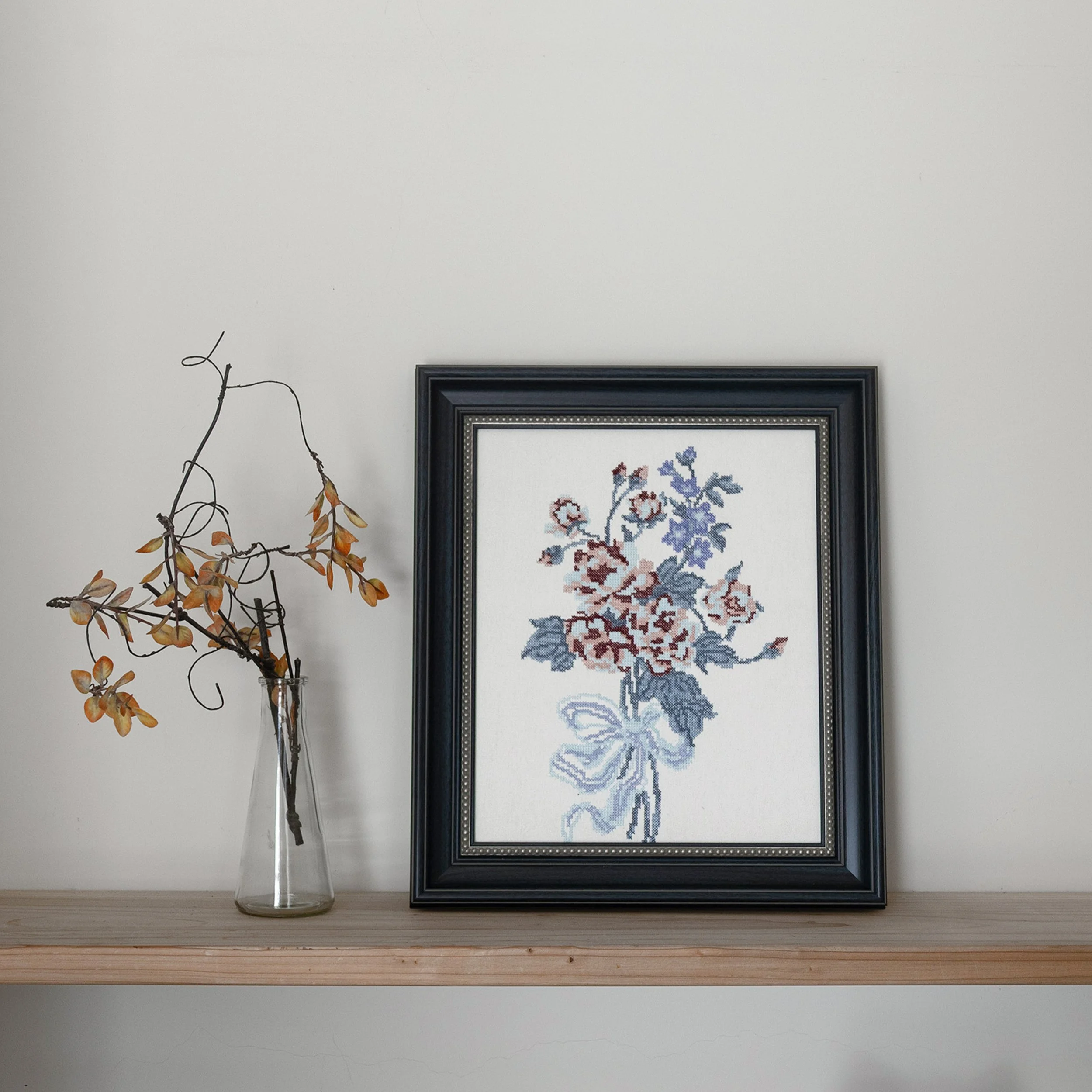 Bouquet embroidered wall art with cross-stitch flowers styled on a wooden shelf in a minimalist interior