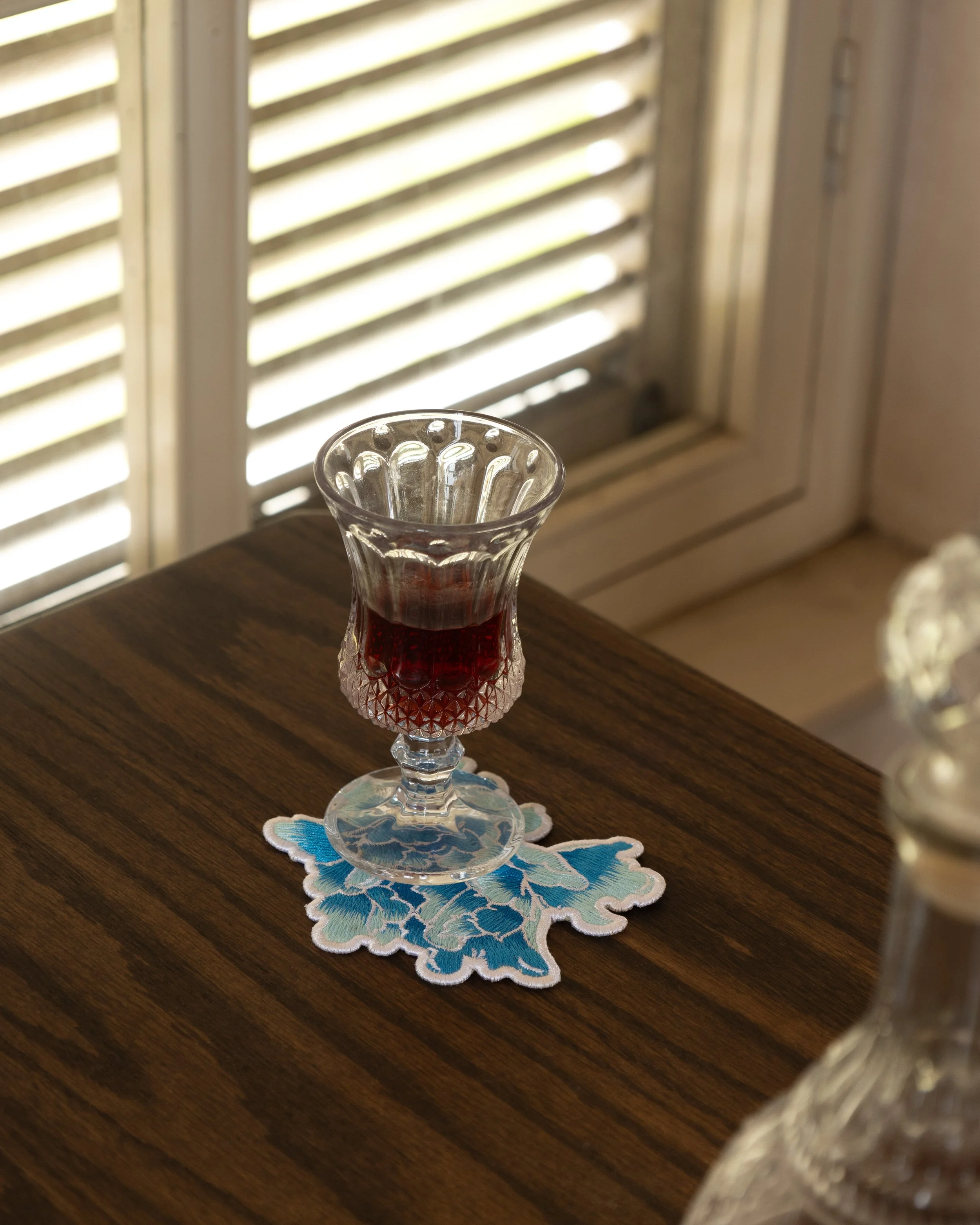 Embroidered coaster from the Etherelle series place on a wooden table with a glass on top.jpg