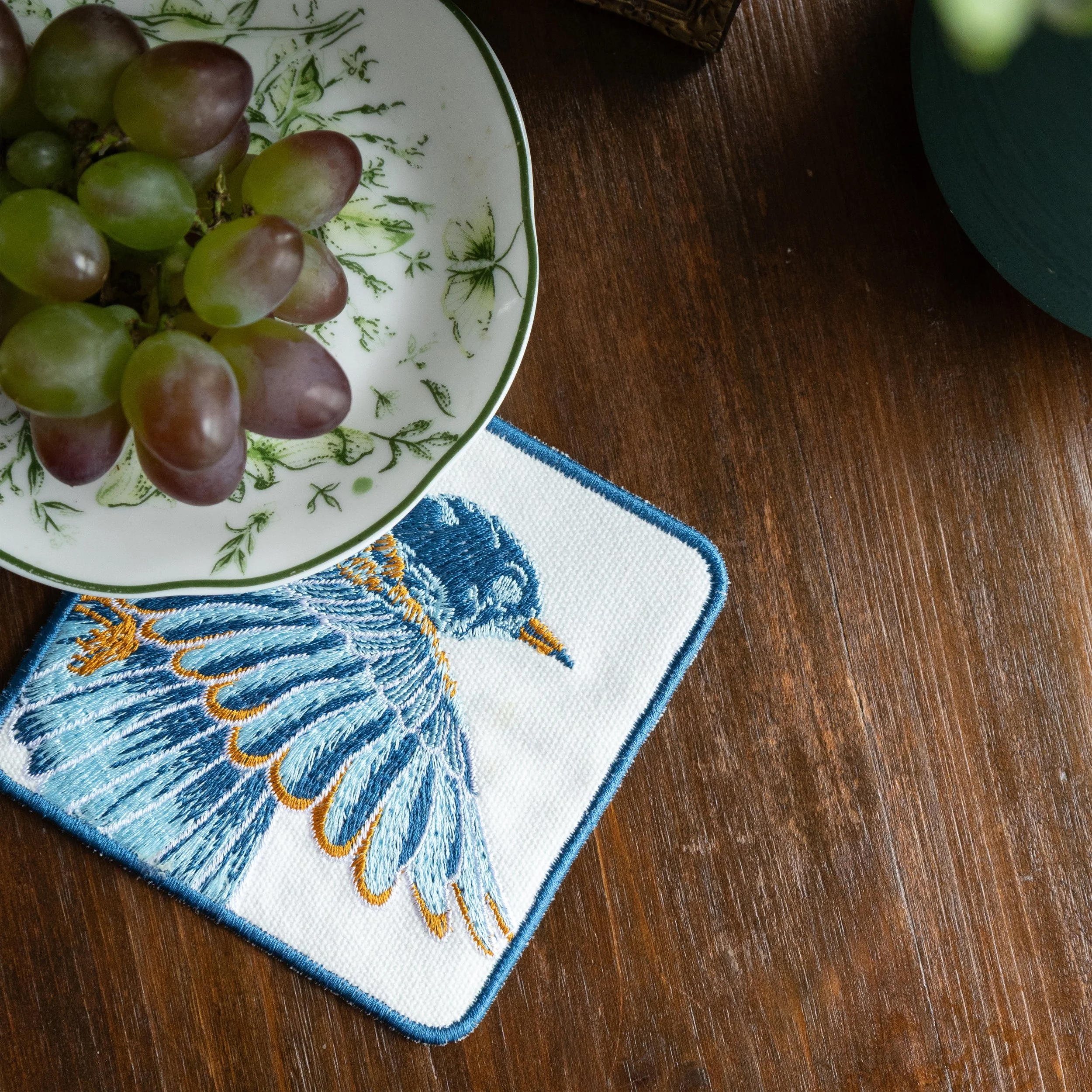 Square coaster with blue bird embroidery placed on a wooden table with a bowl of grapes.jpg