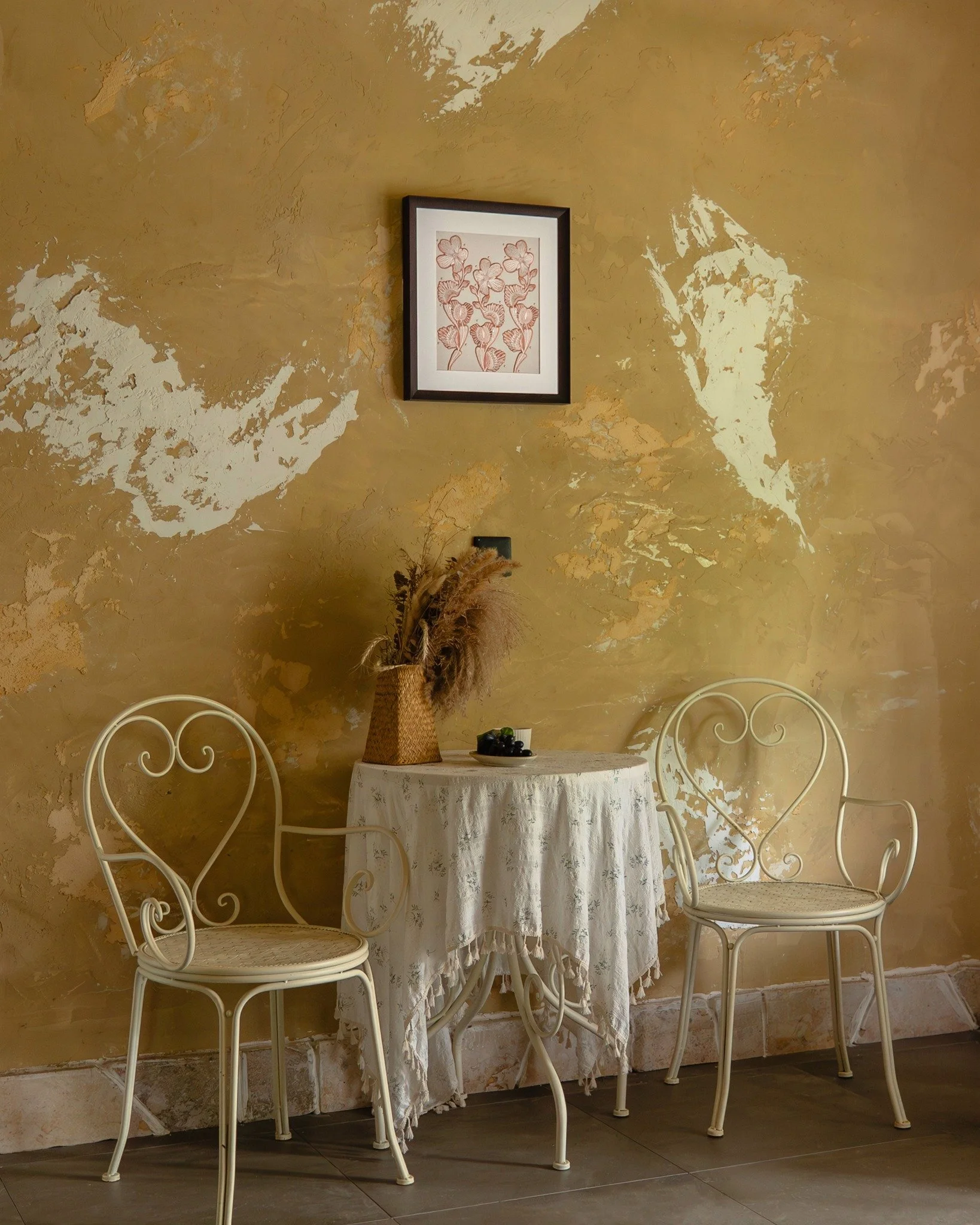 Dionaea embroidery framed art in a warm, vintage countryside corner. 

The space is soft and a little worn in the best way. The textured walls, soft cream metal chairs, and a loosely draped tablecloth already carry a sense of ease.
The piece sits abo