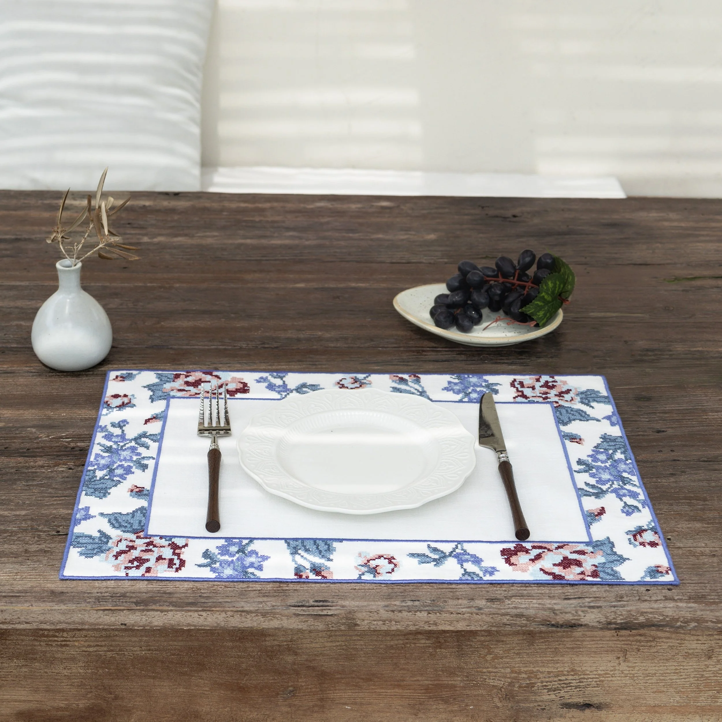 Cross-stitch floral placemat styled on a rustic wooden dining table with white ceramic plate and grapes