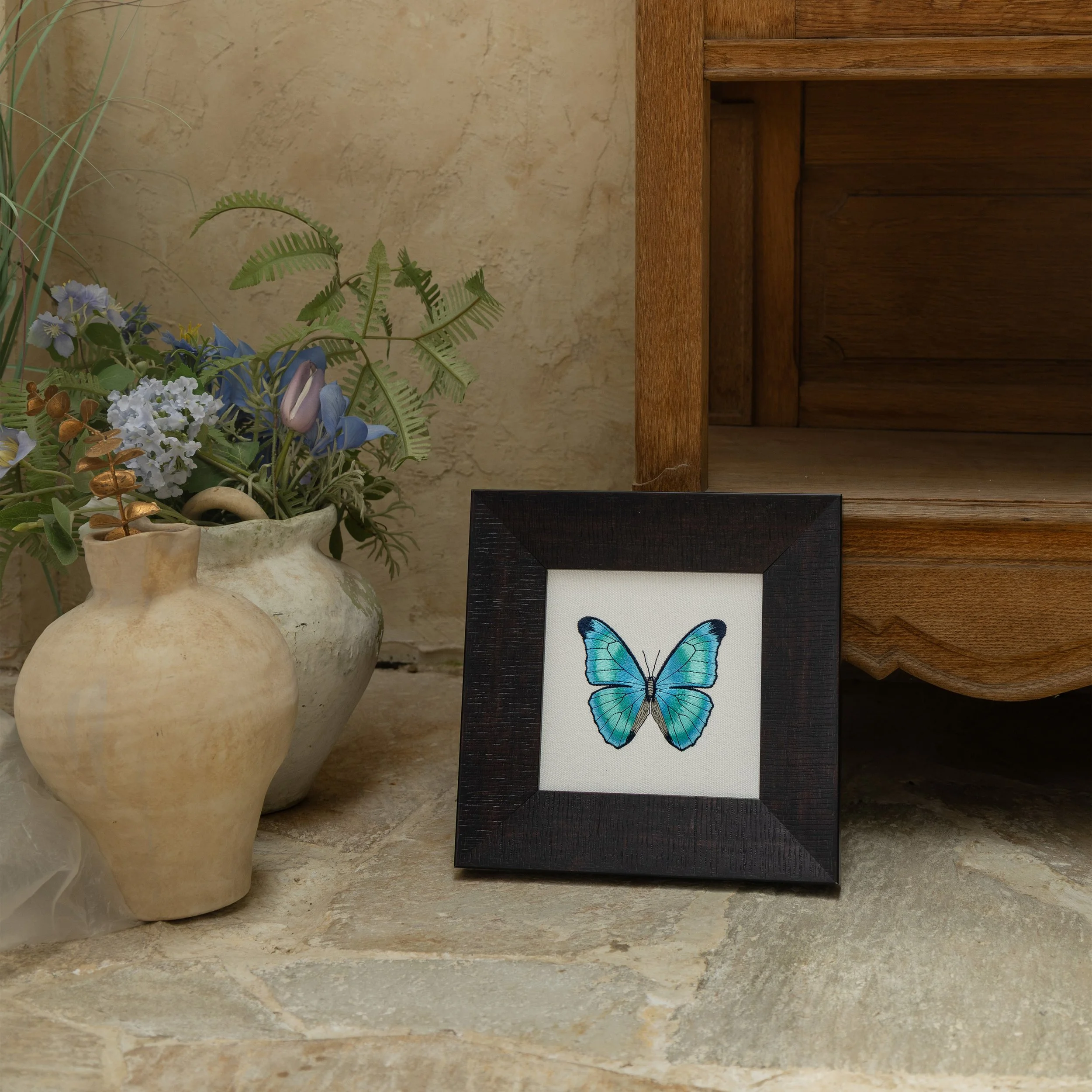 Teal butterfly embroidered wall art styled on a stone floor beside ceramic vessels and greenery.
