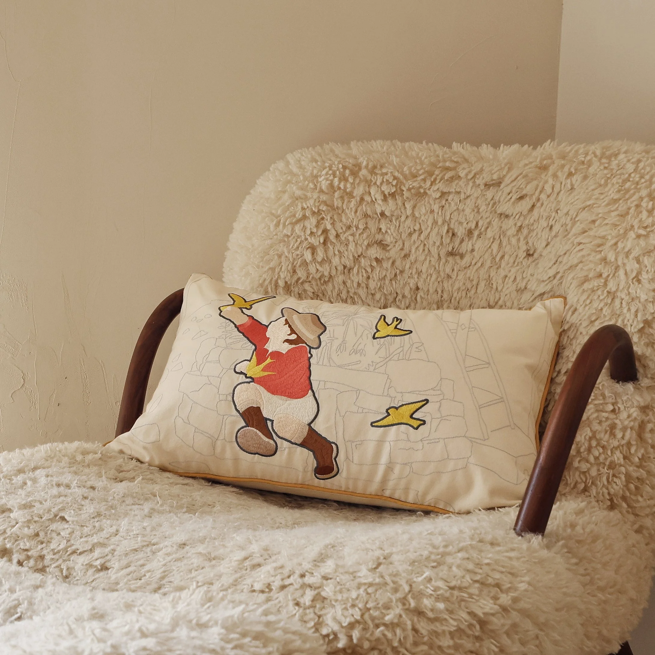 Wild Corner lumbar pillow with playful embroidered child and birds, styled on a cozy sheepskin armchair for a warm, nostalgic touch.