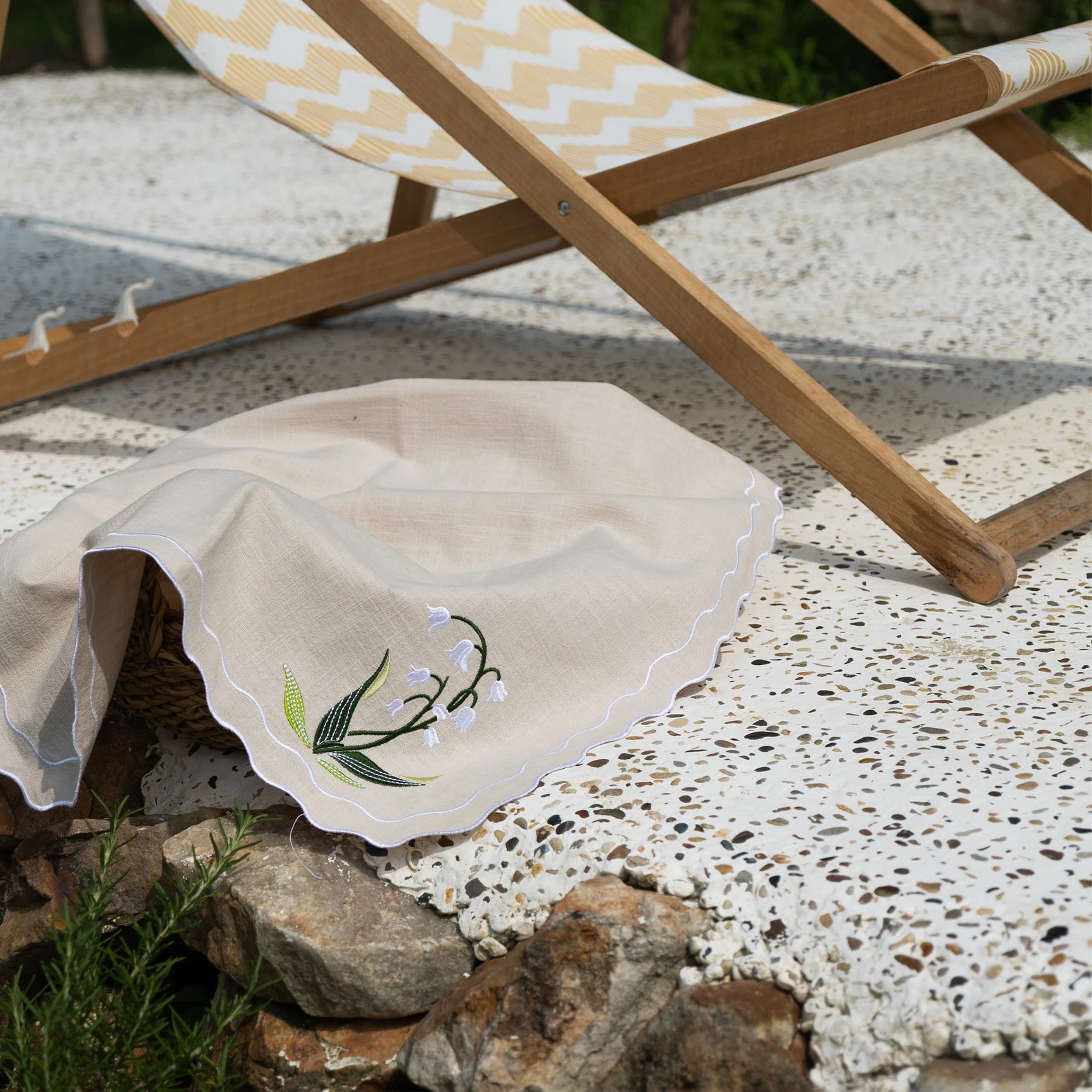 Lily of the valley embroidered napkin draped over a woven basket outdoors, showing scalloped edging and botanical embroidery