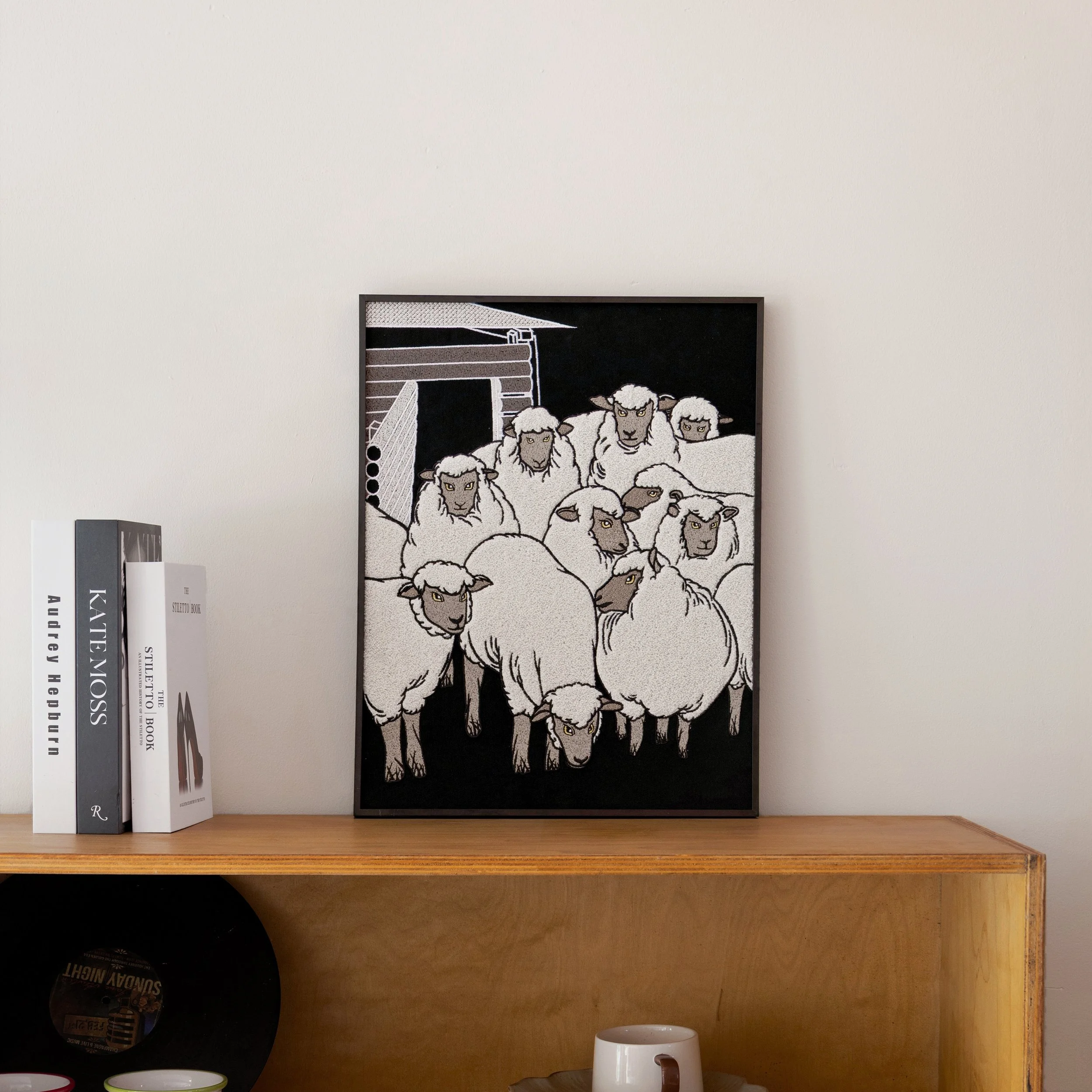 The Flock sheep embroidered framed wall art displayed on a wooden shelf in a modern interior