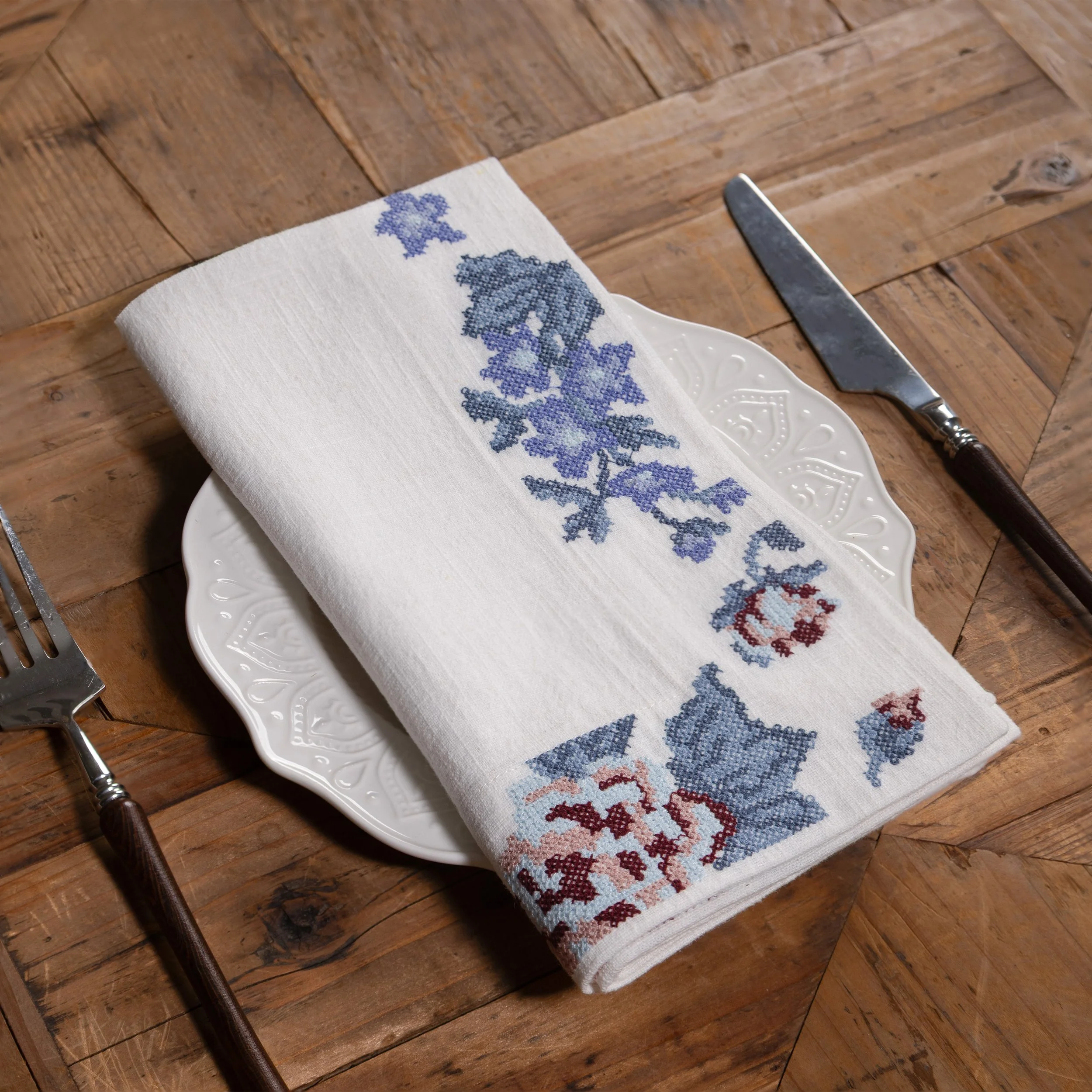 Cross stitch floral embroidered napkin folded on a white ceramic plate atop a rustic wooden table