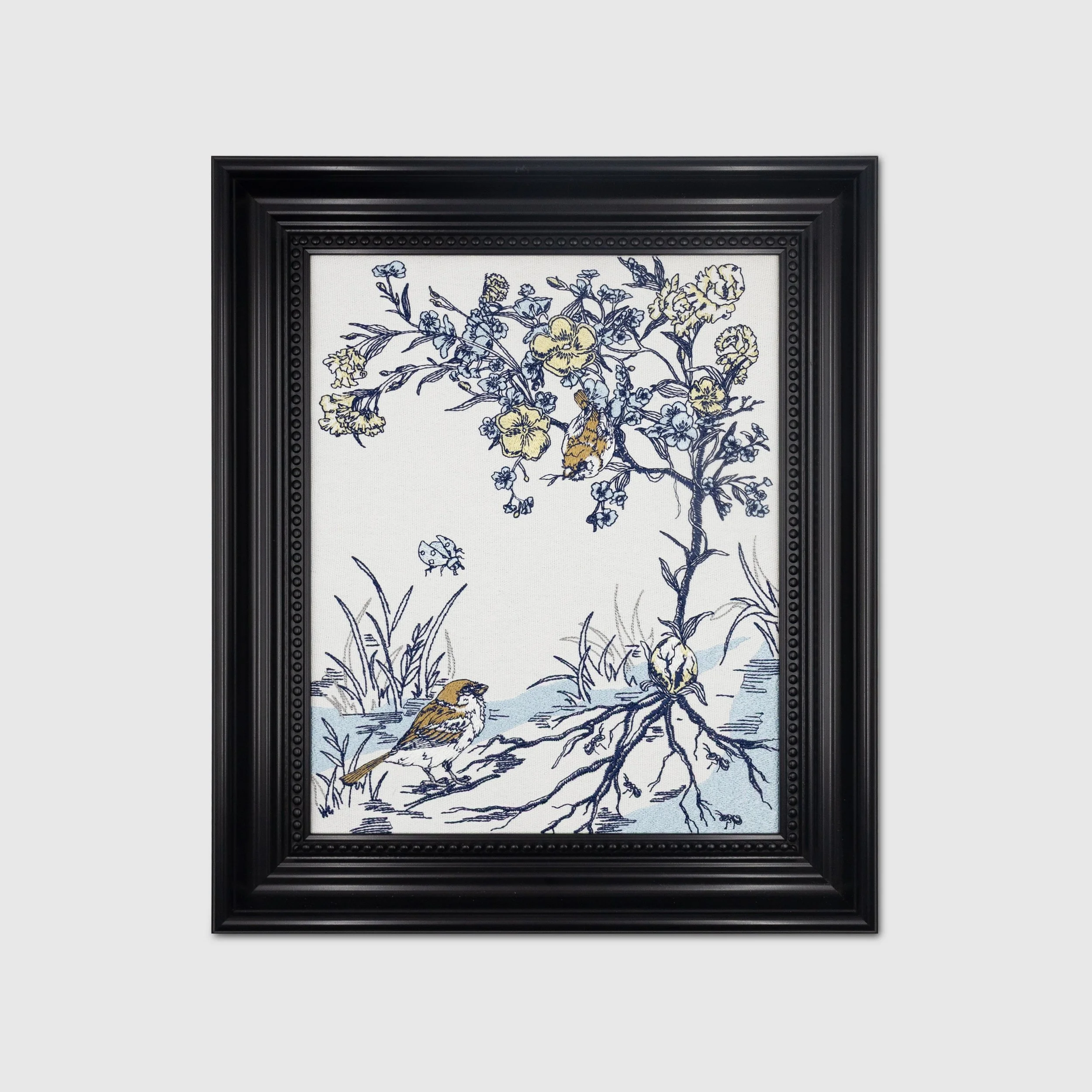 Framed embroidered botanical bird wall art featuring delicate floral branches, small birds, and woodland details on a natural fabric background.