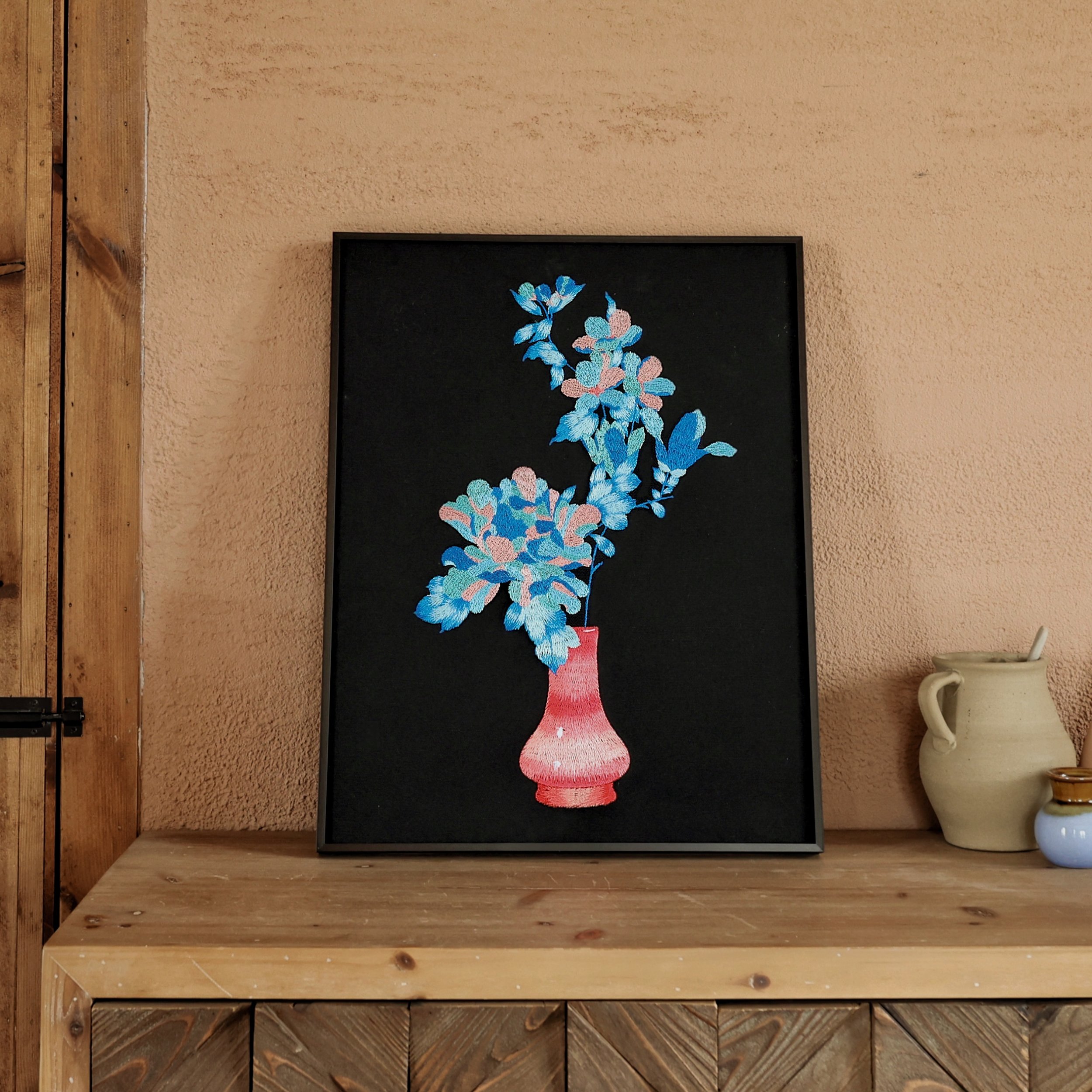 Embroidered blue flowers with pink vase wall art leaning against the wall on a cabinet