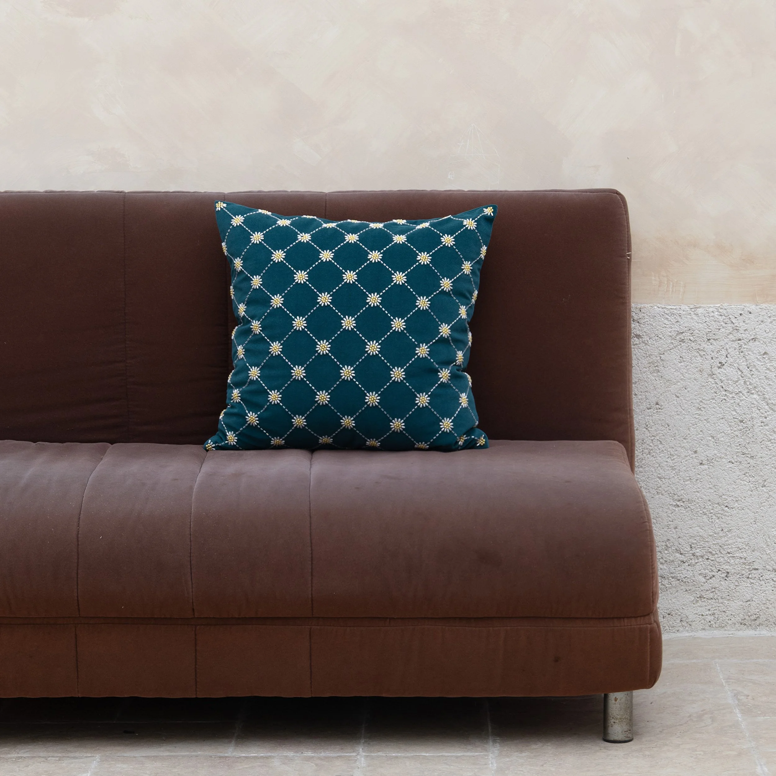 Daisy beaded embroidered throw pillow displayed on a dark brown suede sofa against a textured neutral wall.
