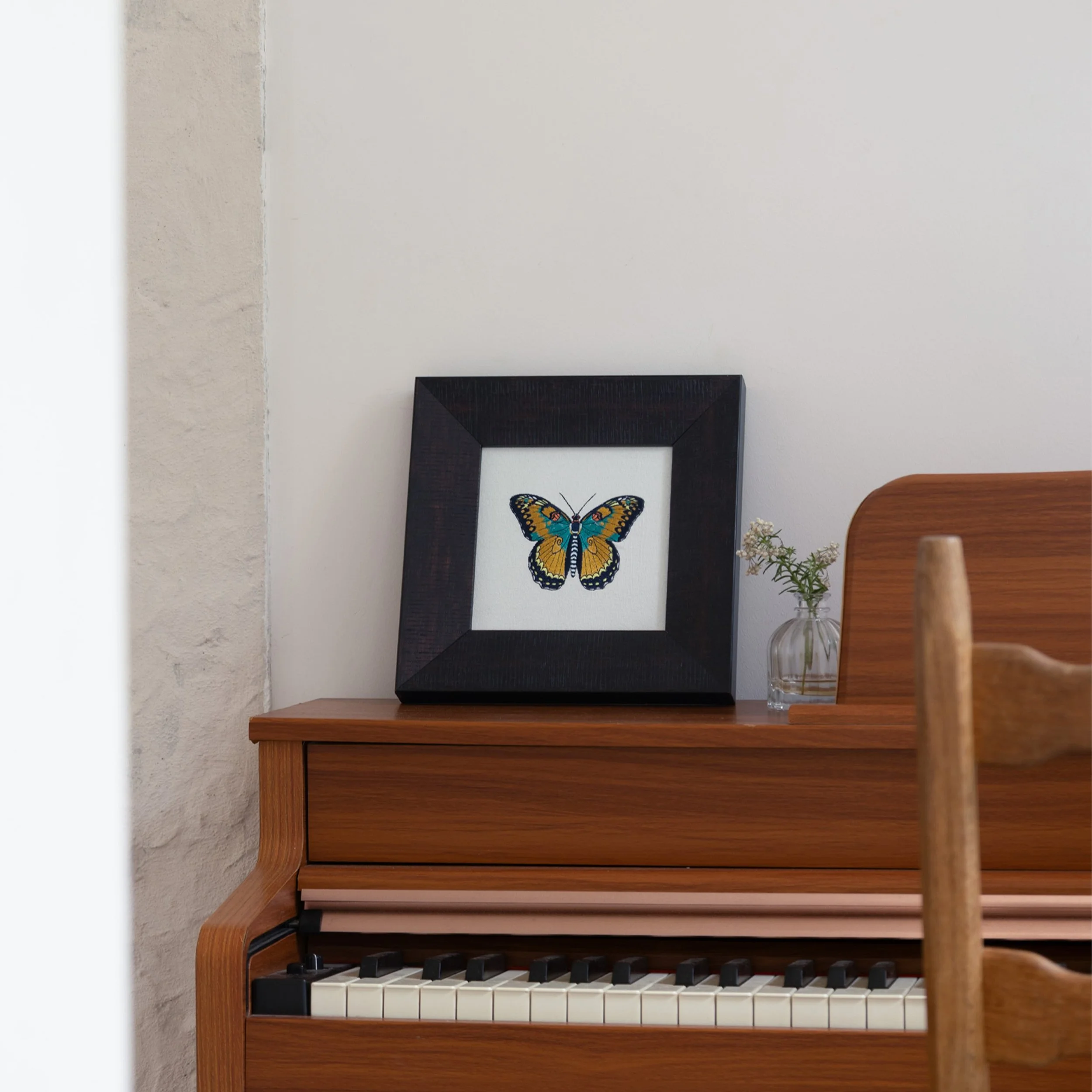Framed yellow and teal butterfly embroidery displayed on a wooden piano with a small glass vase.