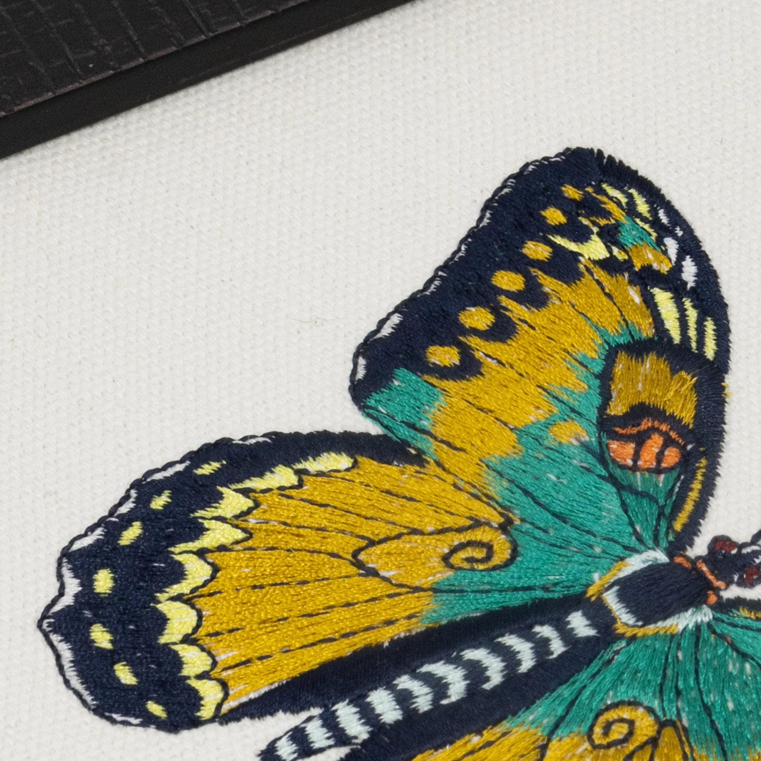 Close-up of Ignisia butterfly embroidery showing refined fine-line stitching in yellow, teal, navy, and orange tones.