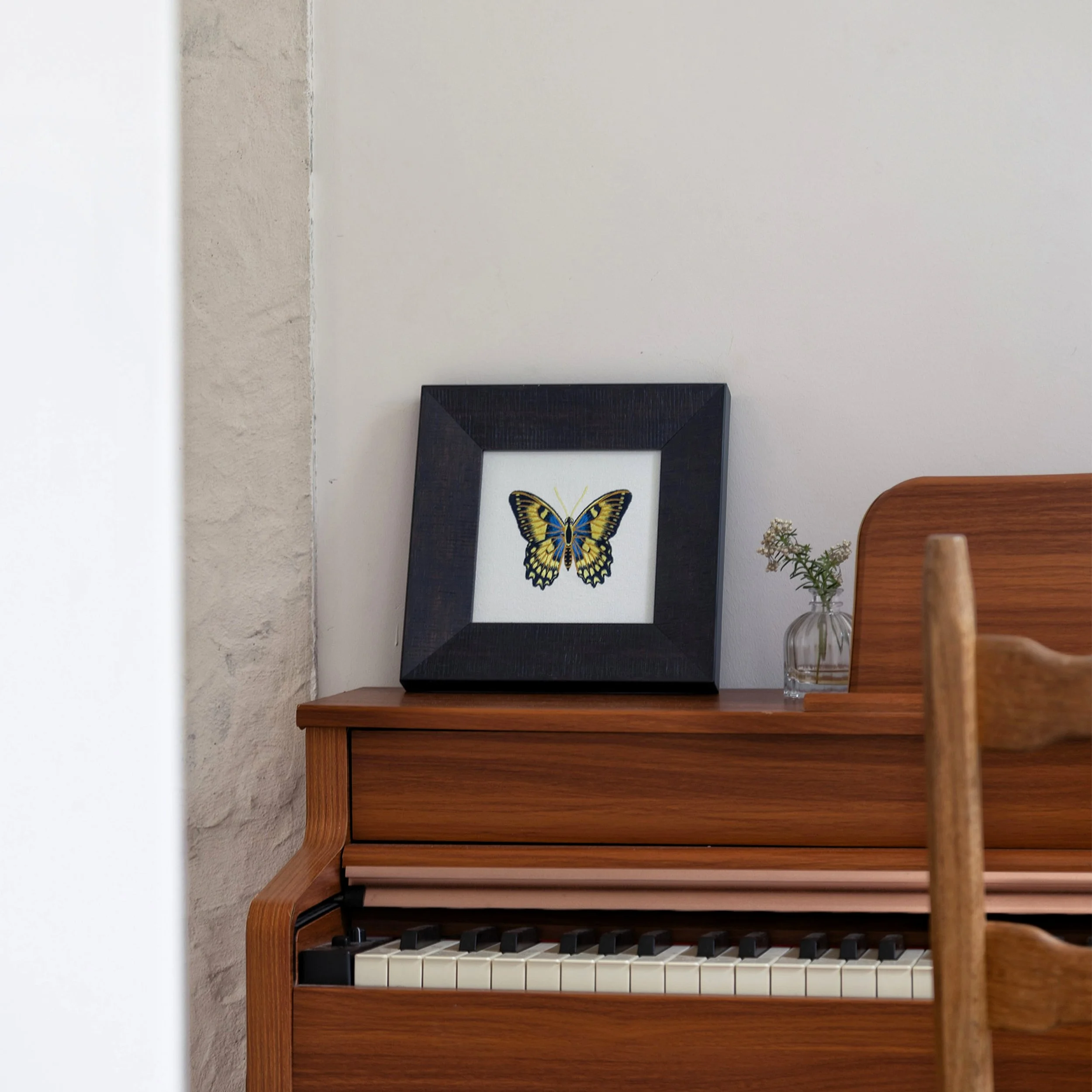 Luminara butterfly embroidered wall art displayed on a wooden piano beside a small glass vase with delicate flowers.