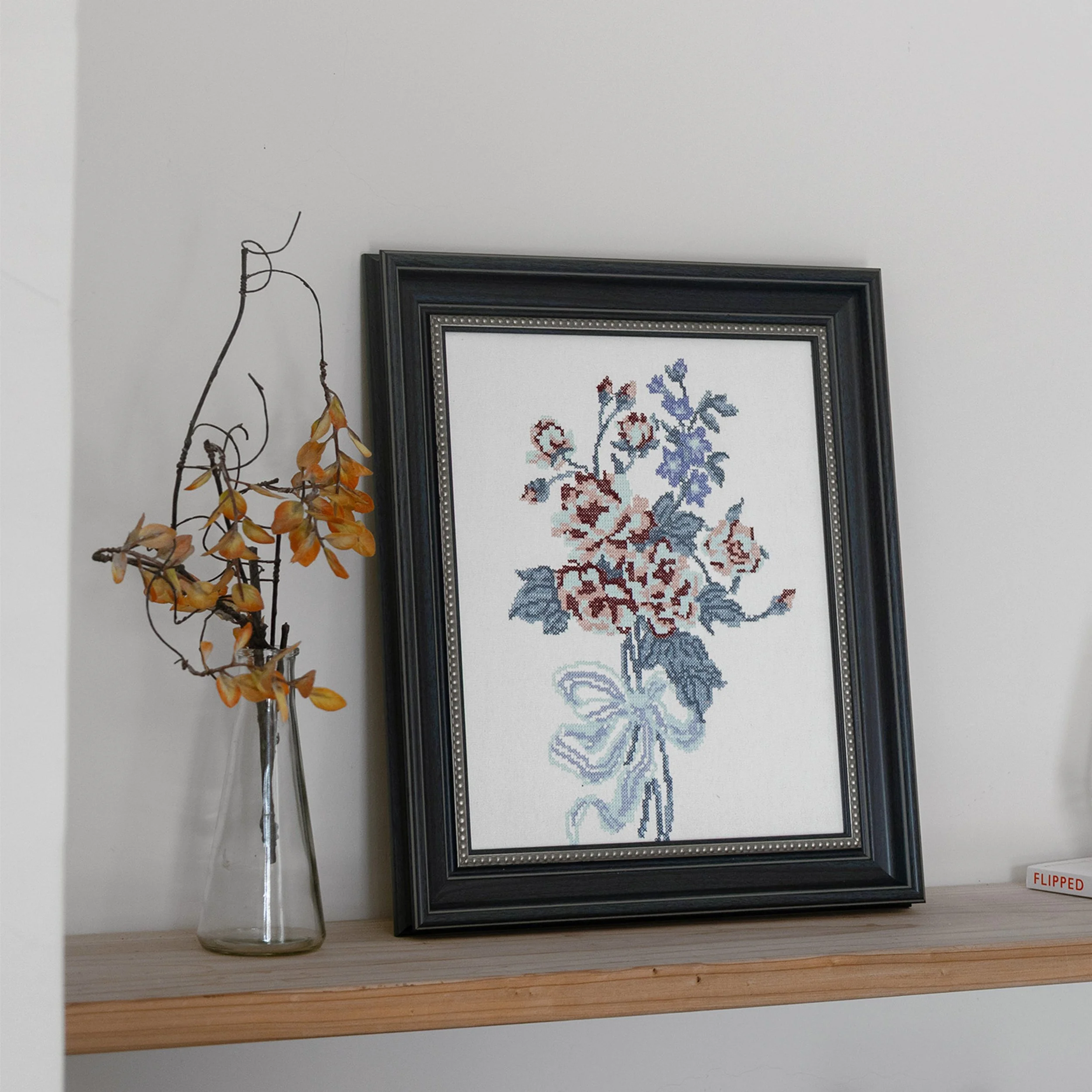 Framed floral cross stitch wall art displayed on a wooden shelf beside a glass vase with dried branches