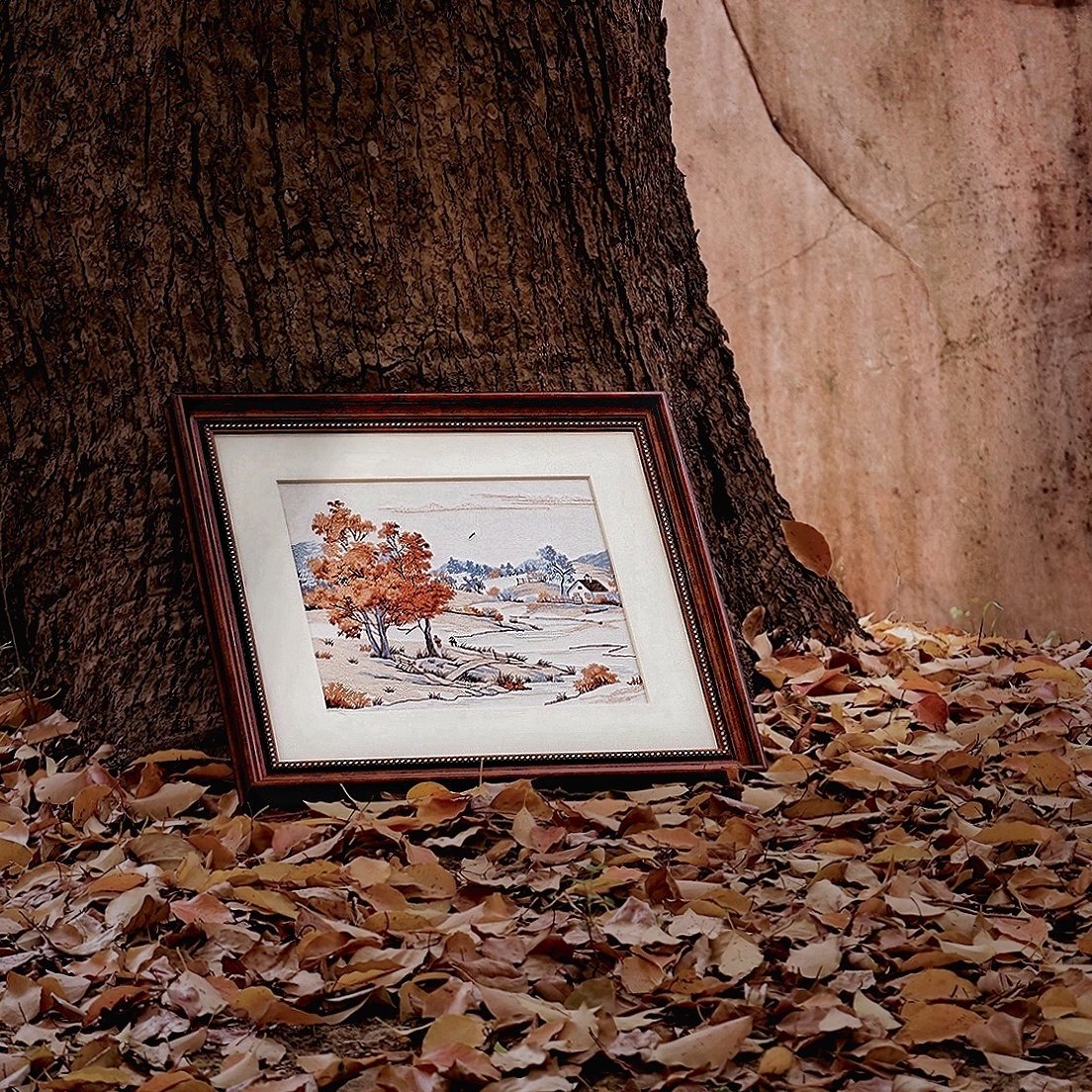Embroidered framed wall art autumn village landscape design leaning against tree trunk in fallen leaves