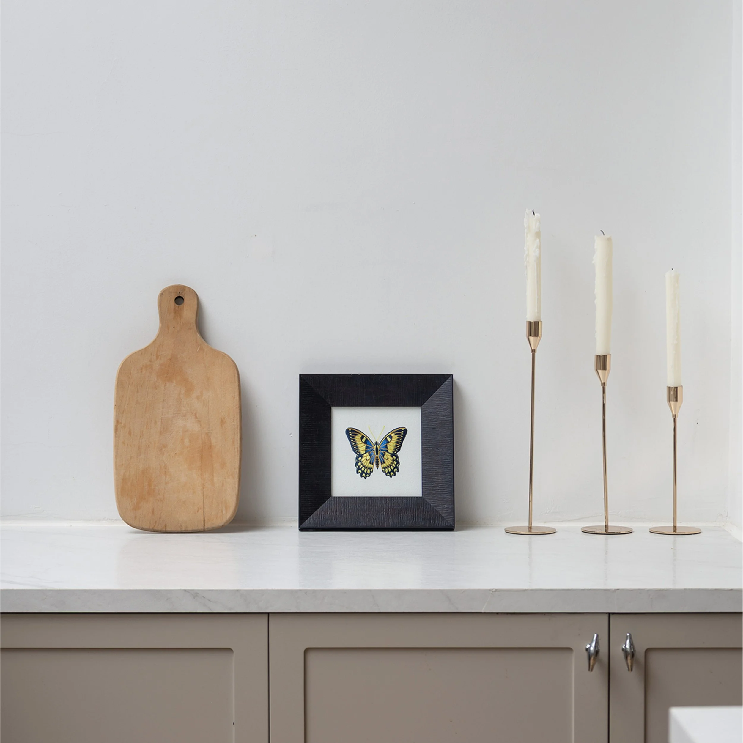 Framed blue and yellow butterfly embroidery styled on a white countertop alongside a wooden cutting board and candle holders.