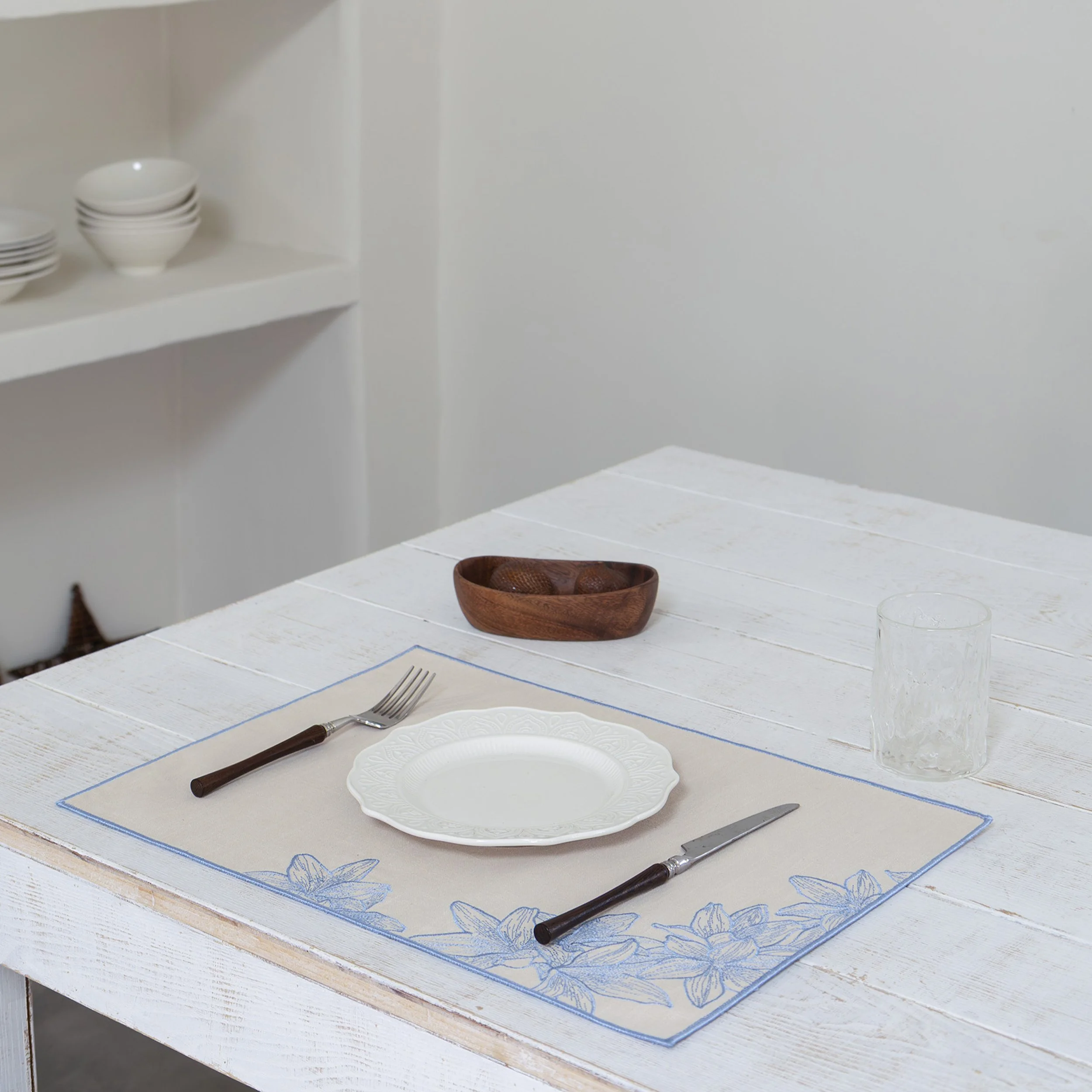 Floral embroidered placemat with blue lily motif on a white wooden dining table with modern tableware